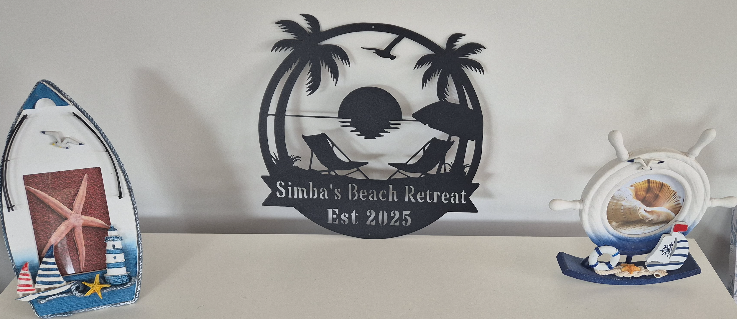 Decorative beach-themed items including a surfboard with a starfish and lighthouse picture, a black metal sign with palm trees, beach chairs, a sunset, and the text 'Simba's Beach Retreat Est 2025', and a nautical figurine with a conch shell and sailboat design.