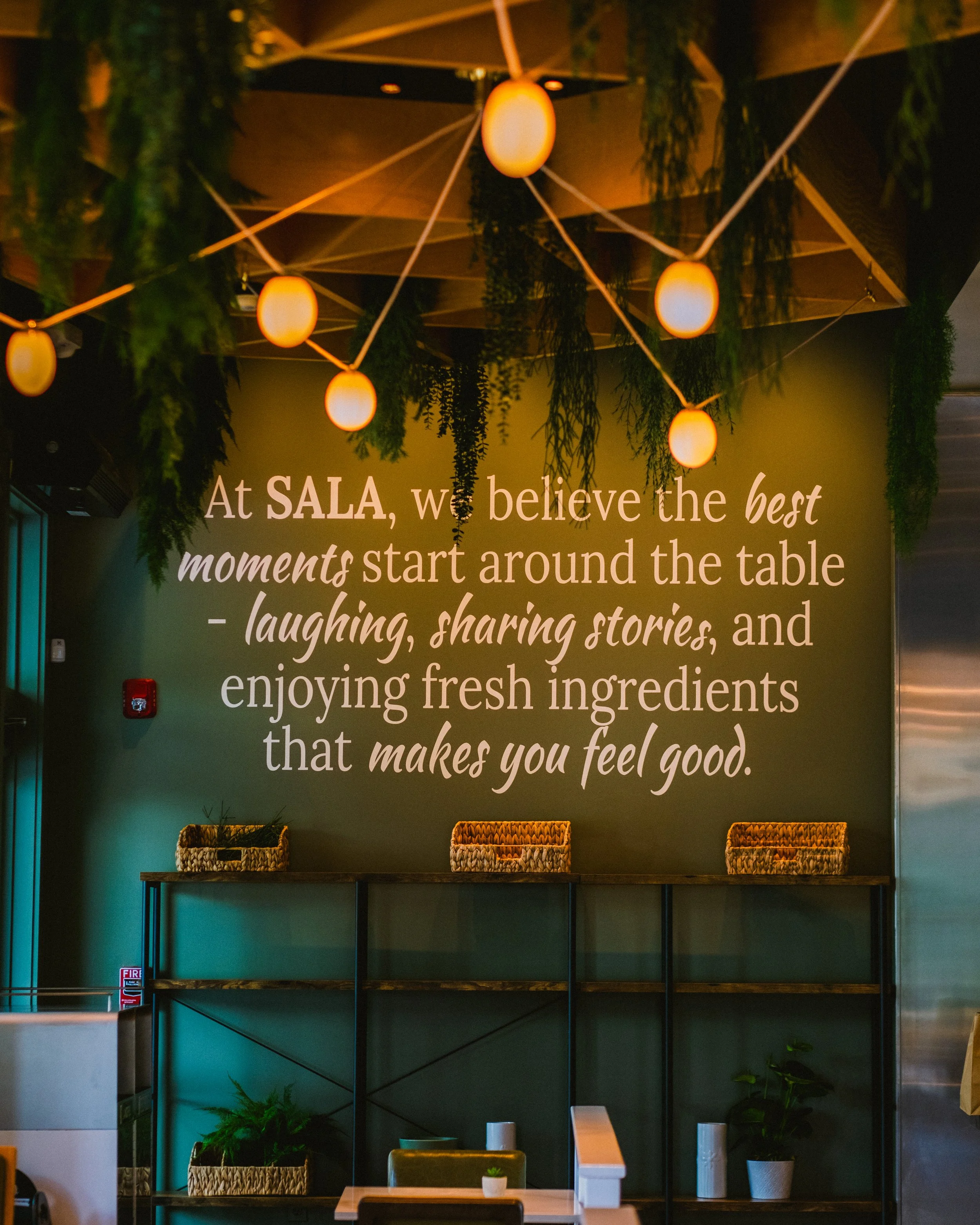 Interior of a restaurant with hanging string lights, plants, a wall with an inspirational quote.
