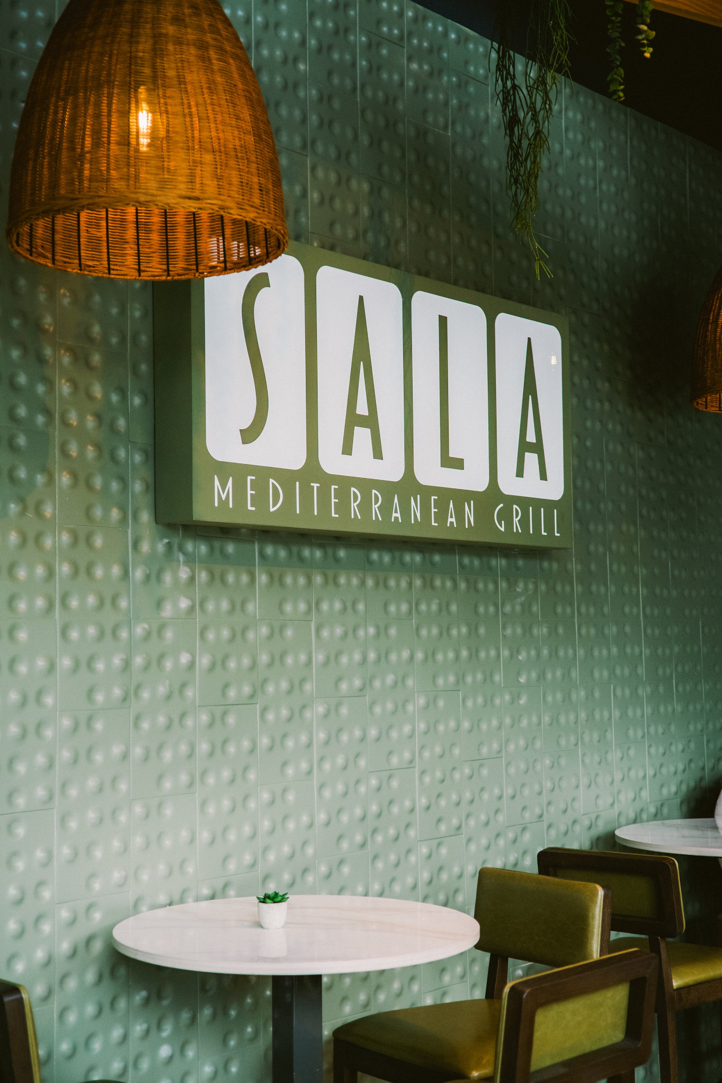 Interior of Sala Mediterranean Grill with a sign on a green textured wall, featuring hanging wicker light fixtures, a small white table with a potted plant, and olive green chairs.