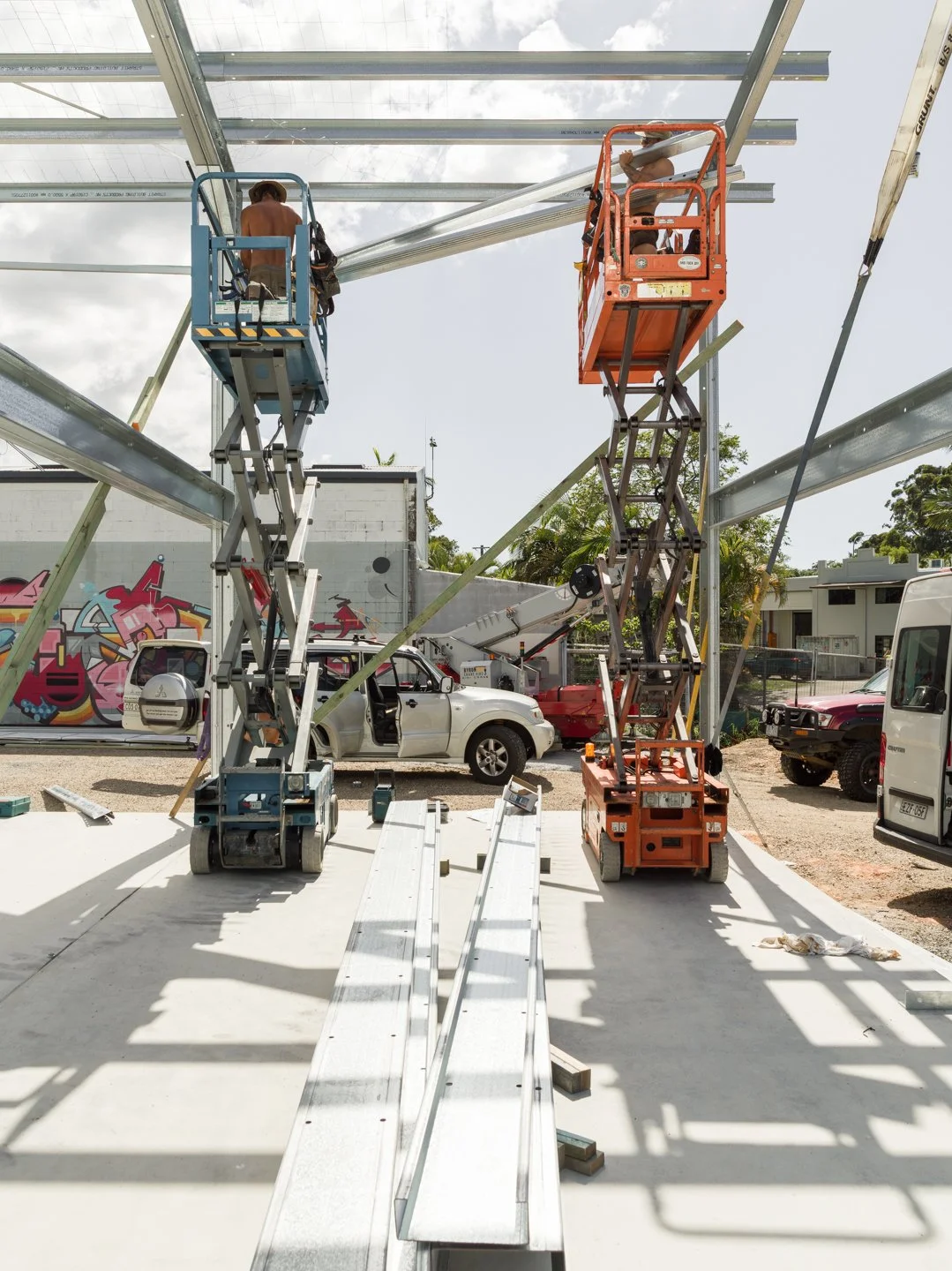 Construction workers are installing a metal framework structure using scissor lifts and a cherry picker, with a white truck and graffiti mural visible in the background.