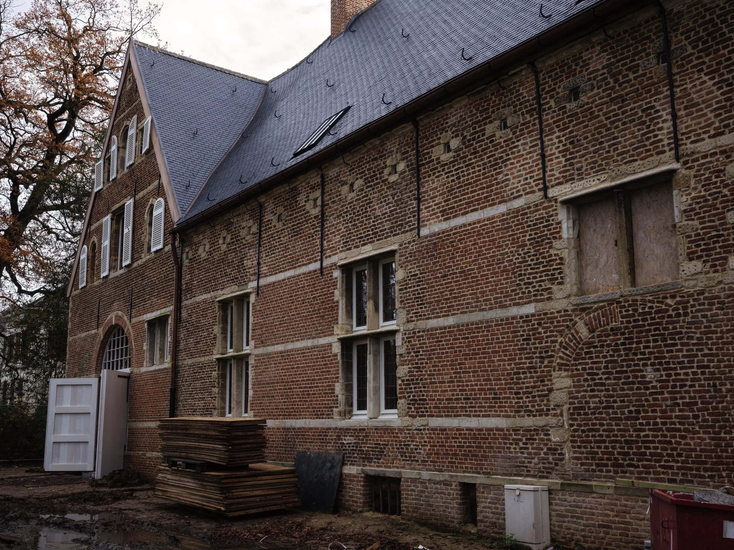 A brick building under renovation with boarded-up windows, construction materials stacked outside, and a white door slightly open, surrounded by fall trees.