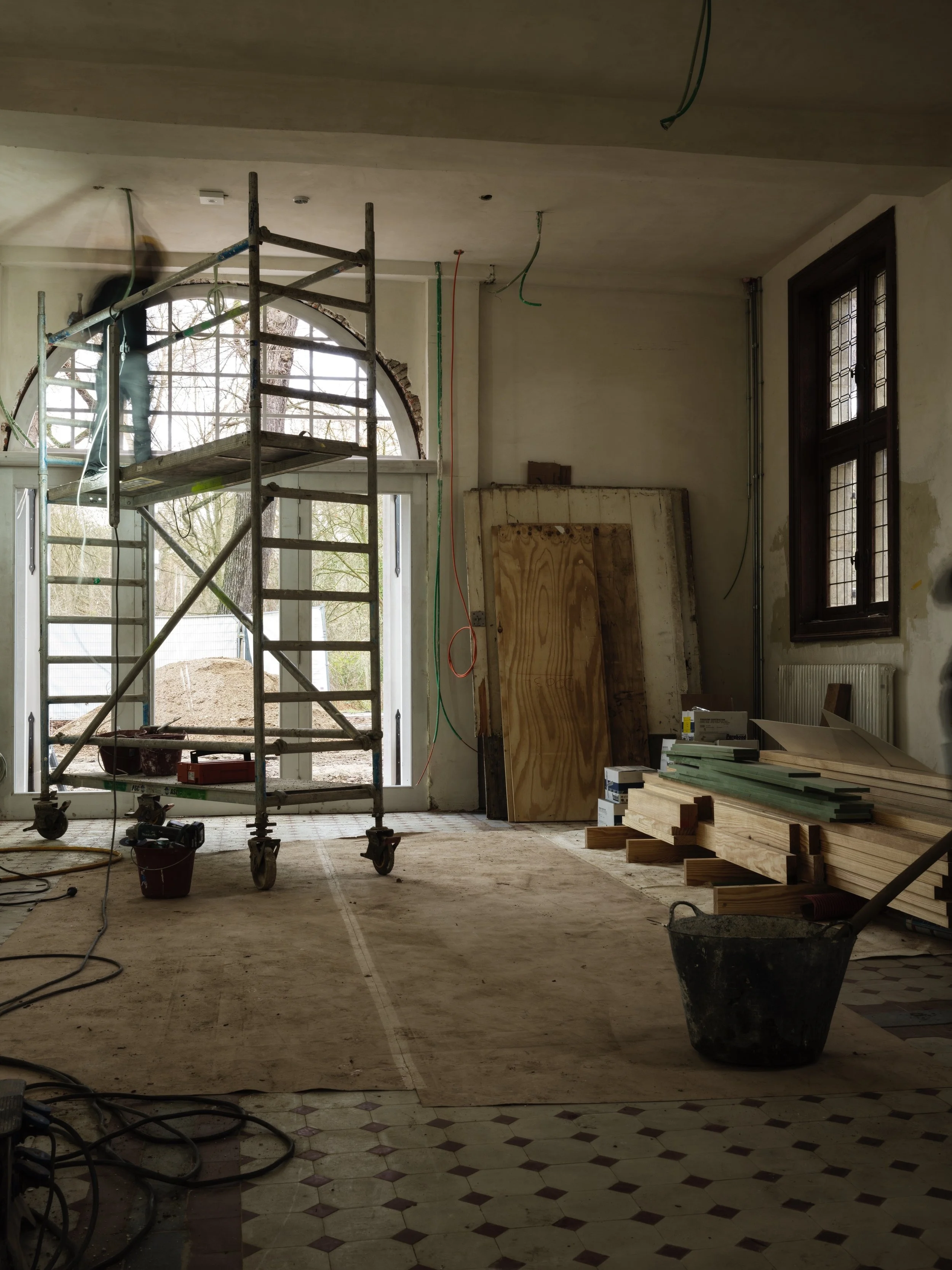 Interior of a room under renovation with construction tools, wooden planks, scaffolding, and a worker on a scaffold near a large arched window.