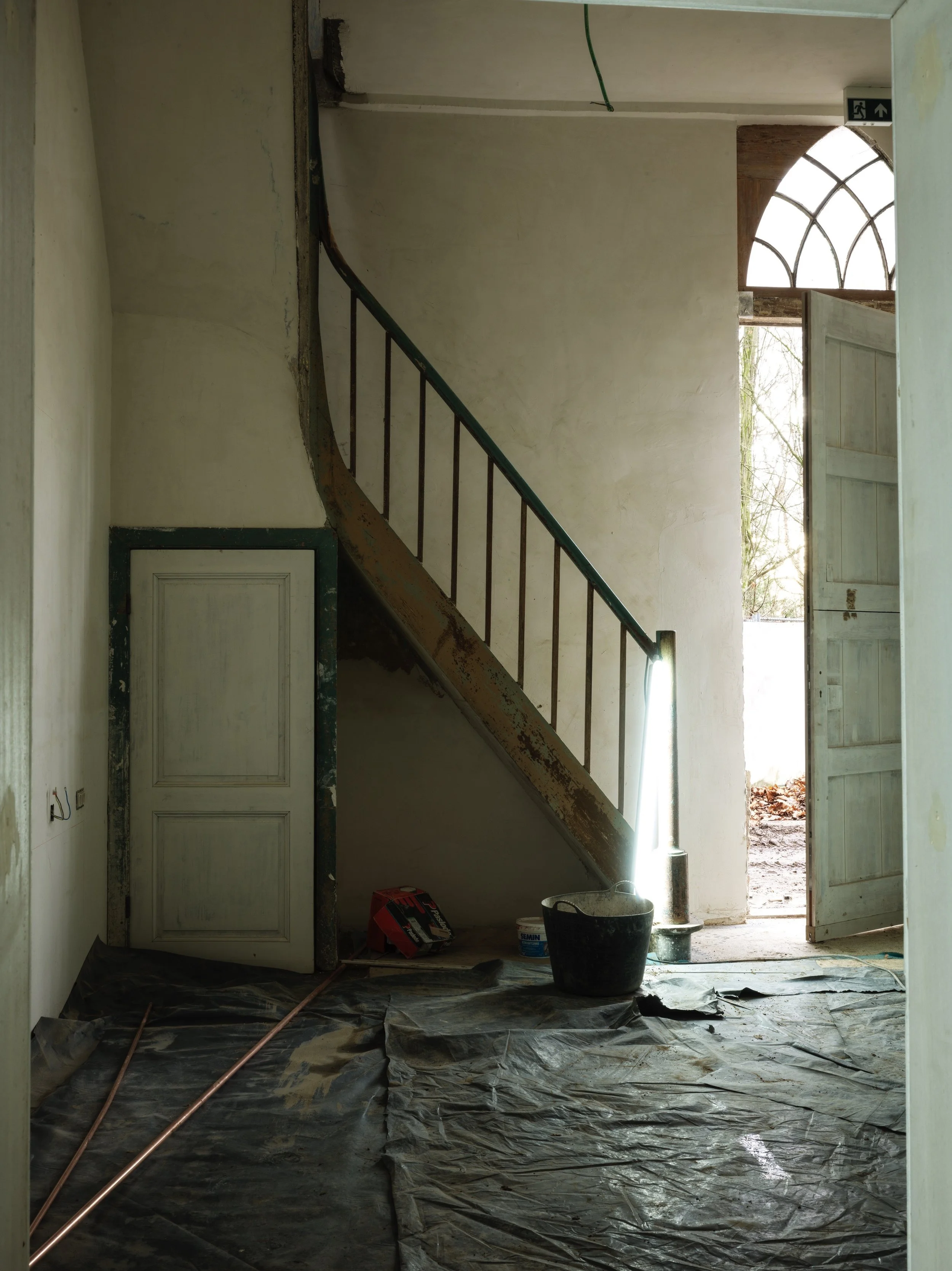 Interior of a house under renovation with an open door and a staircase with a rusty railing, construction tools, buckets, and a plastic tarp on the floor.
