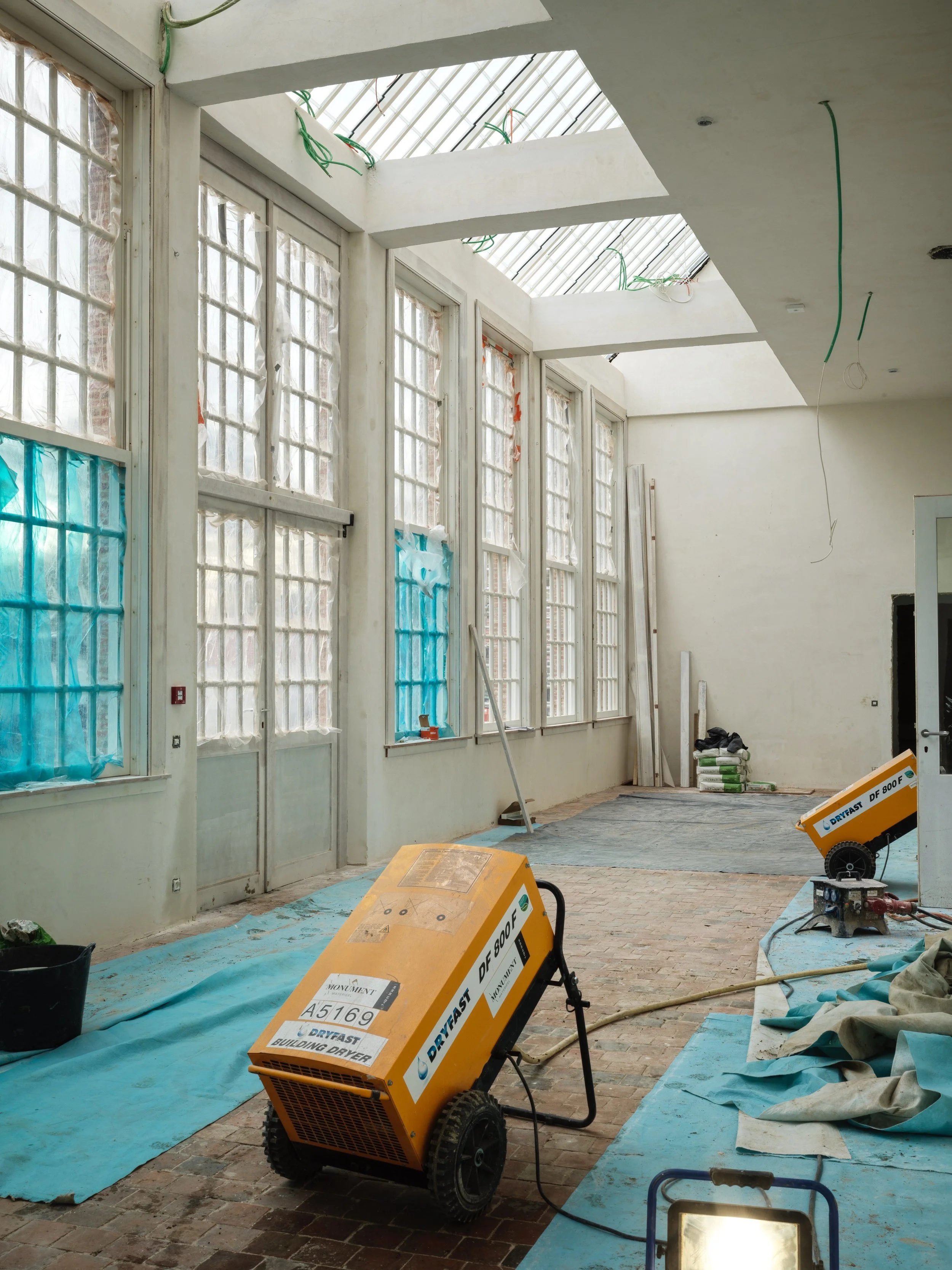 Interior view of a room under construction with large windows, some covered with blue plastic, and construction equipment on the floor.
