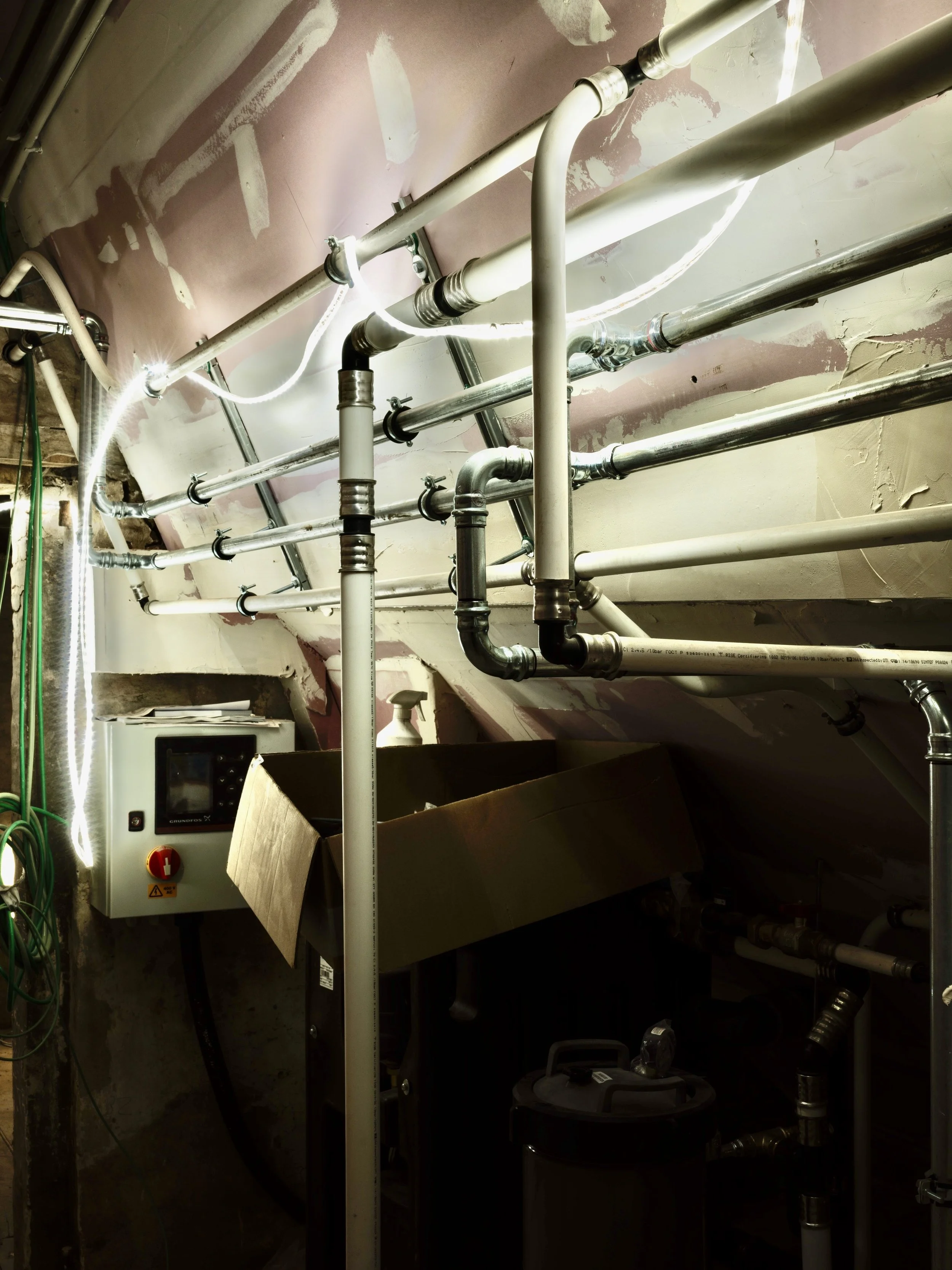 Industrial plumbing system with various pipes, valves, and a control panel in a basement or utility room.