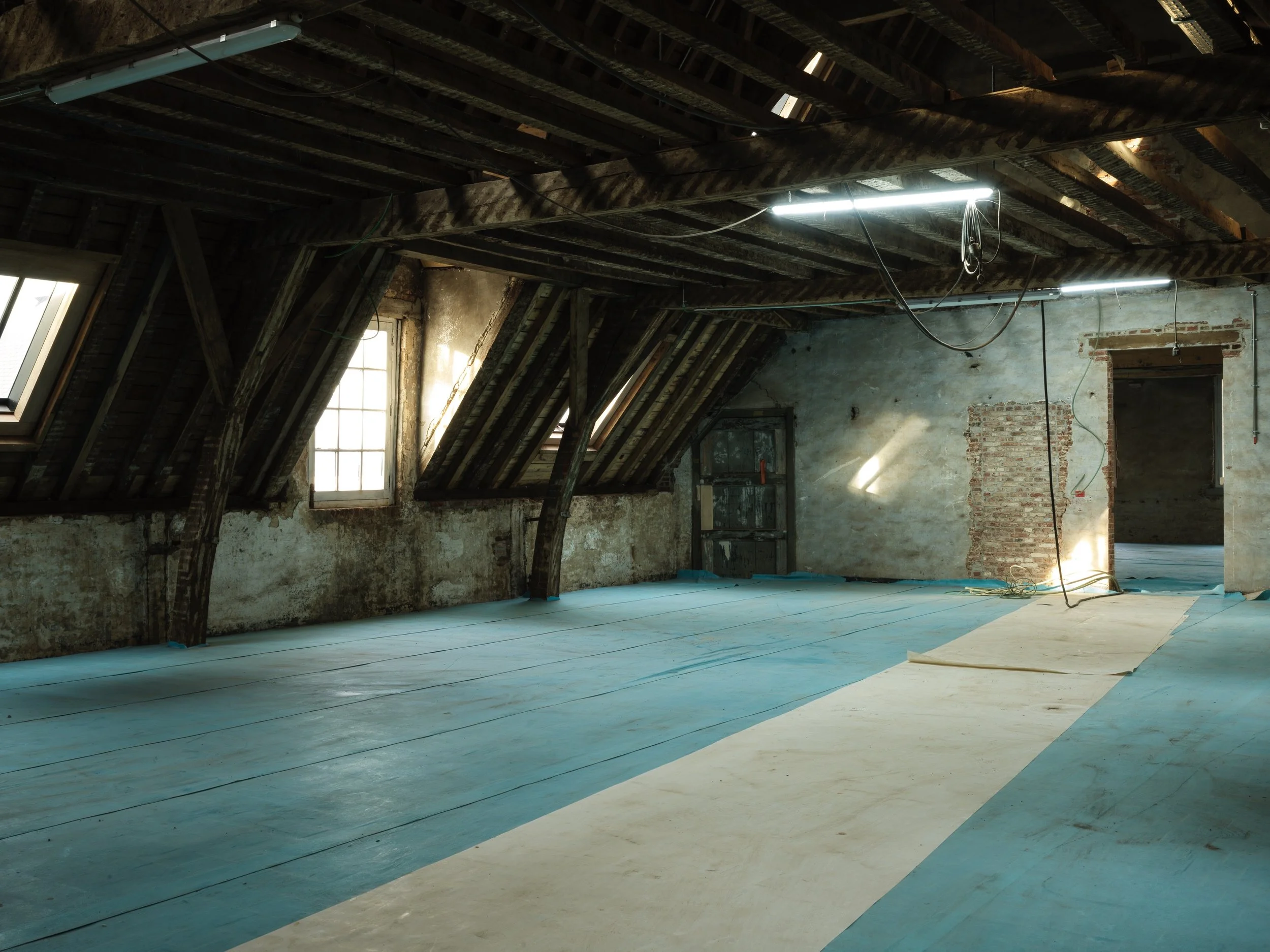 An attic under renovation with exposed wooden beams, distressed walls, and skylights, covered with blue and beige protective flooring.