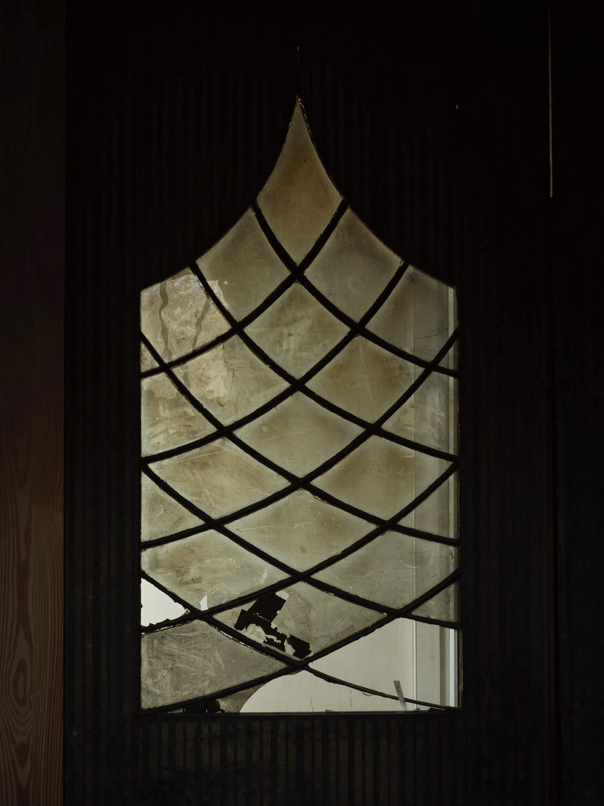 A stained glass window with a decorative black metal grid pattern, viewed from inside a building, with a silhouette of a saw and other tools outside.