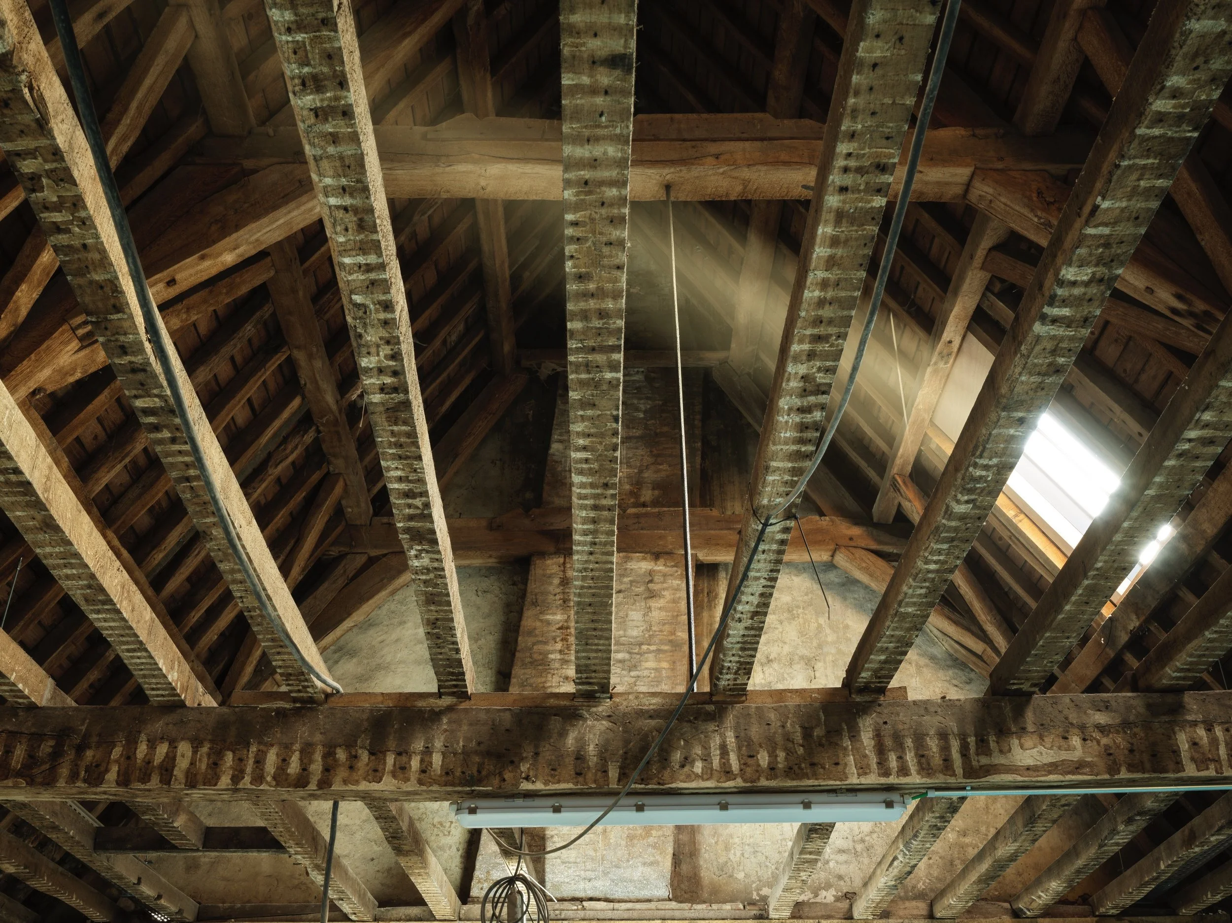 Interior view of a building with exposed wooden beams and rafters in the ceiling. Some electrical wiring and a fluorescent light fixture are visible. Sunlight streams through a small opening in the roof.