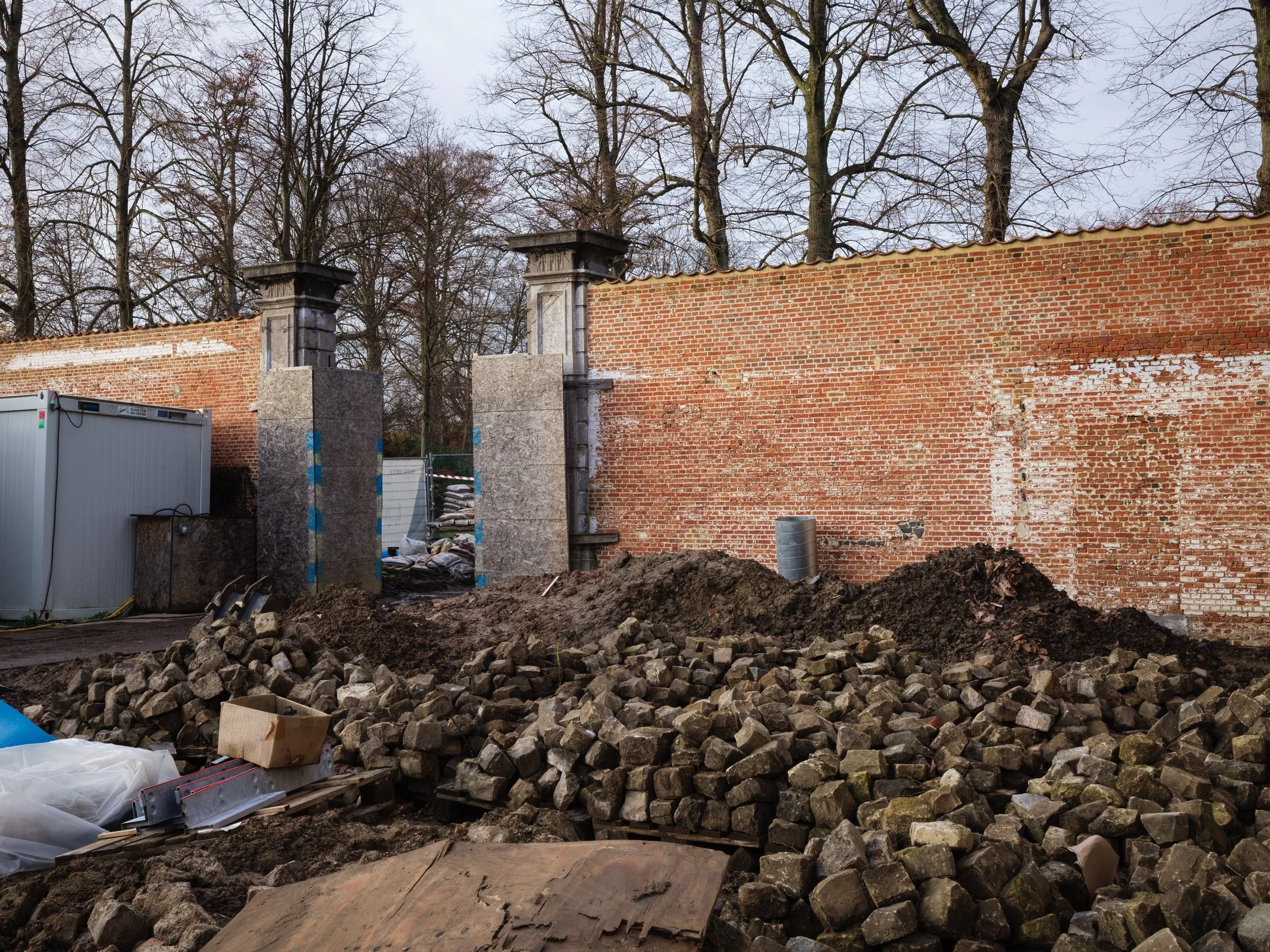 Construction site with piles of bricks and dirt in front of a brick wall and trees in the background.