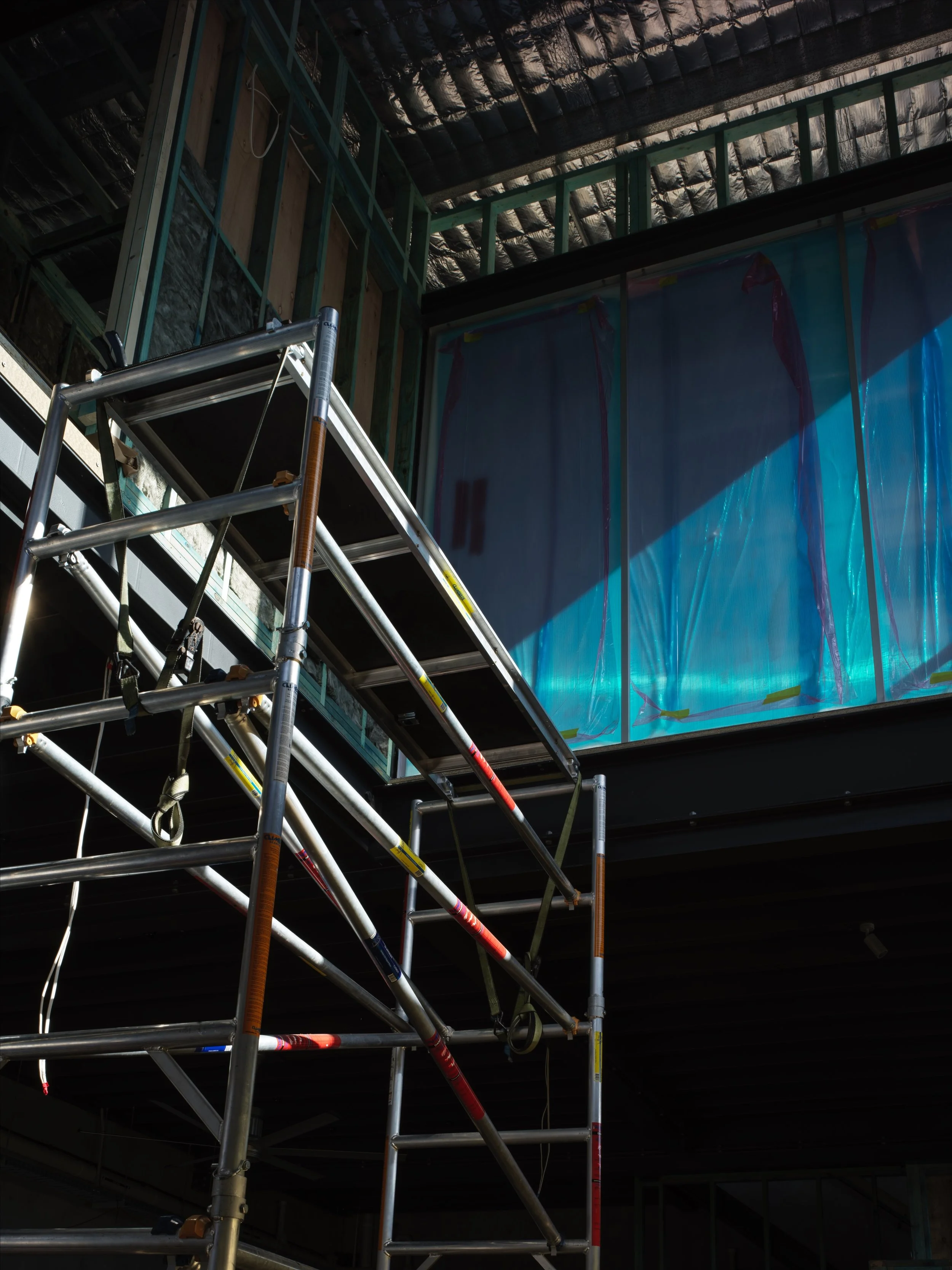 Construction site interior with metal scaffolding and partially framed walls, large plastic sheet covering part of the wall, and exposed ceiling insulation.