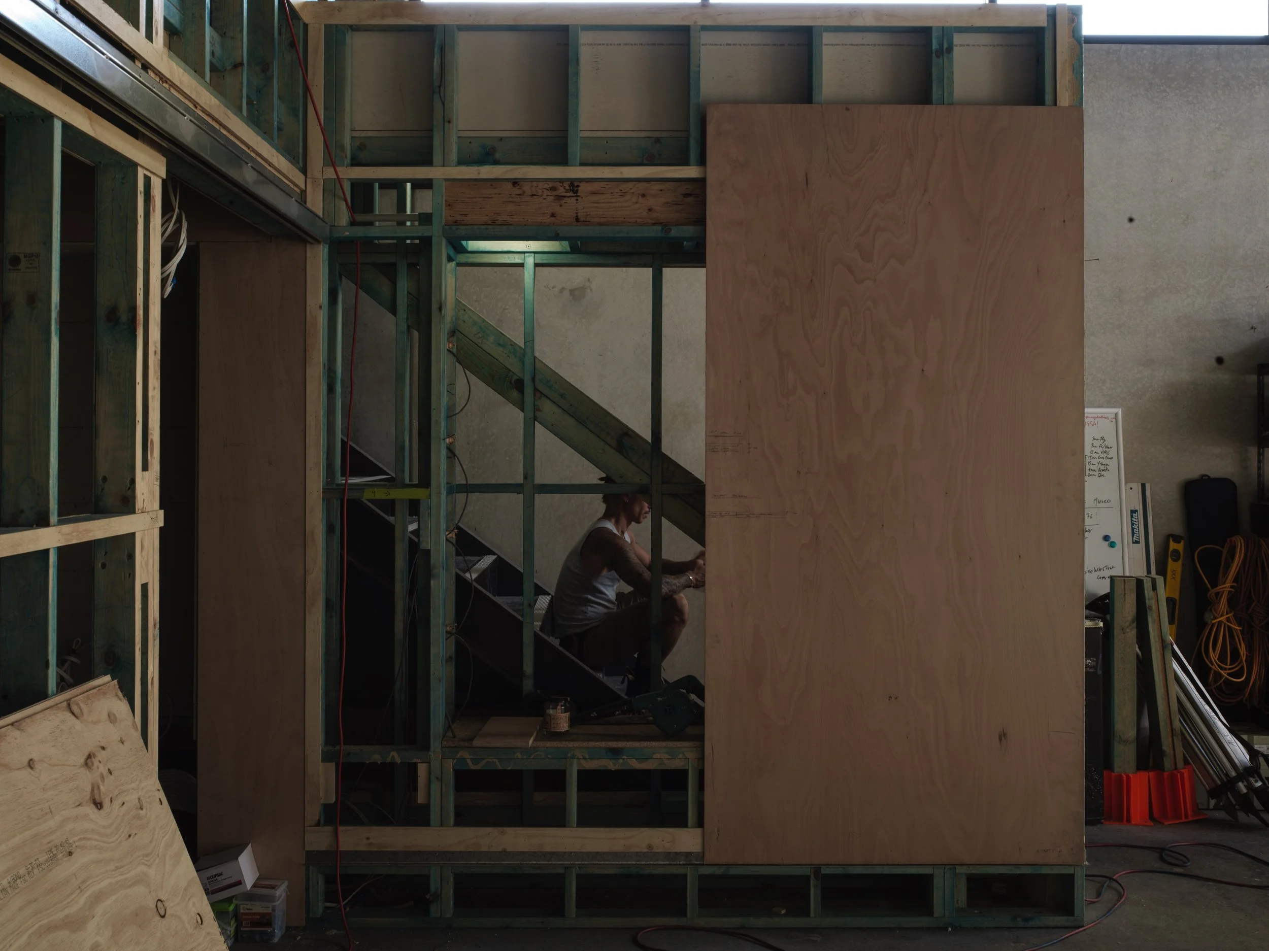 Construction site with wooden framing and stairs, worker sitting on stairs, tools and equipment around.