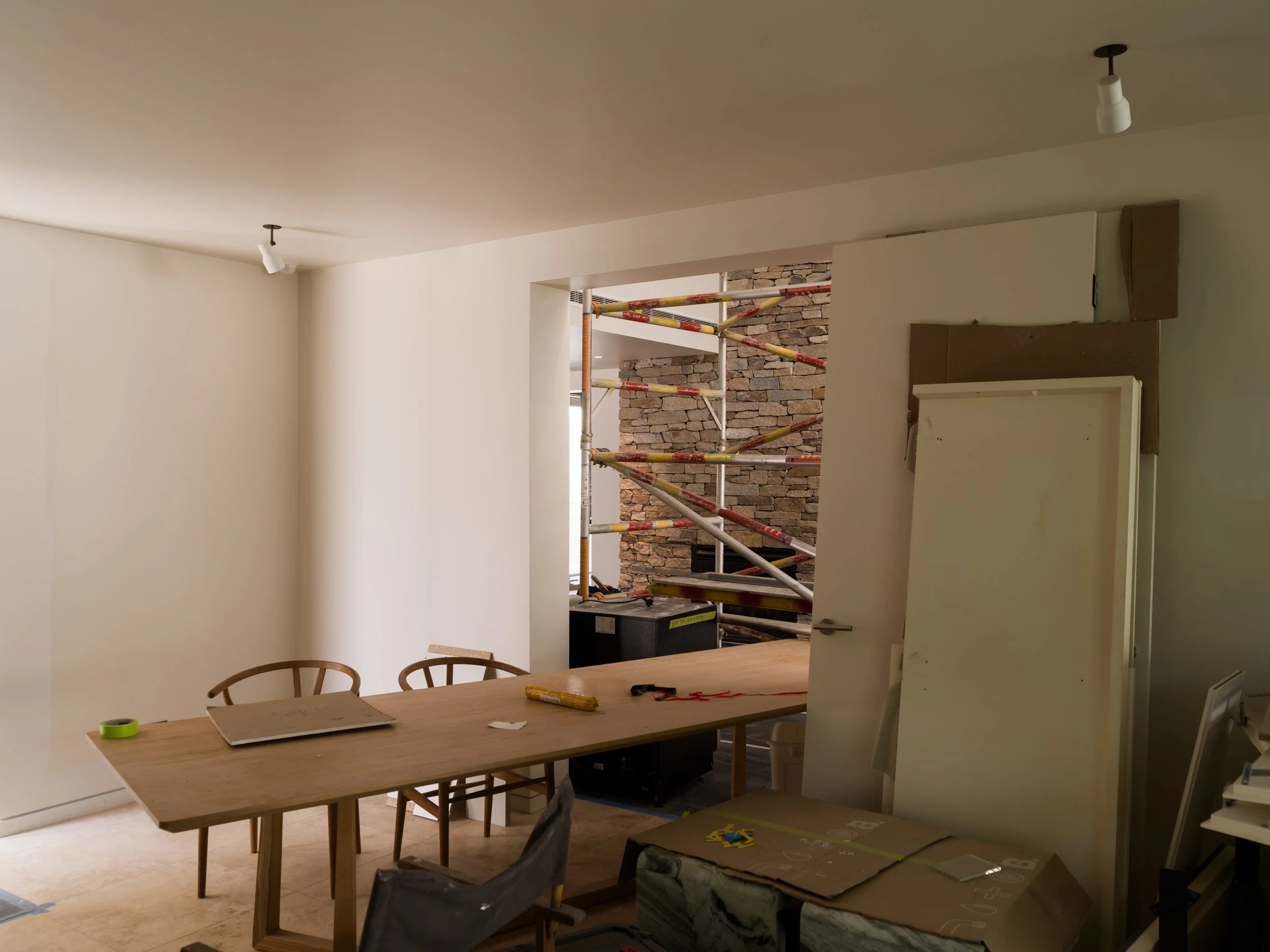 Interior of a room under renovation with a wooden dining table and chairs, construction materials, and scaffolding visible through an open doorway leading to an exposed brick wall.