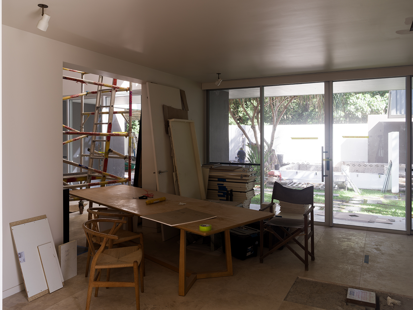 Interior of a room under construction with a wooden table, chairs, construction materials, and large sliding glass doors leading to a backyard with trees and equipment.