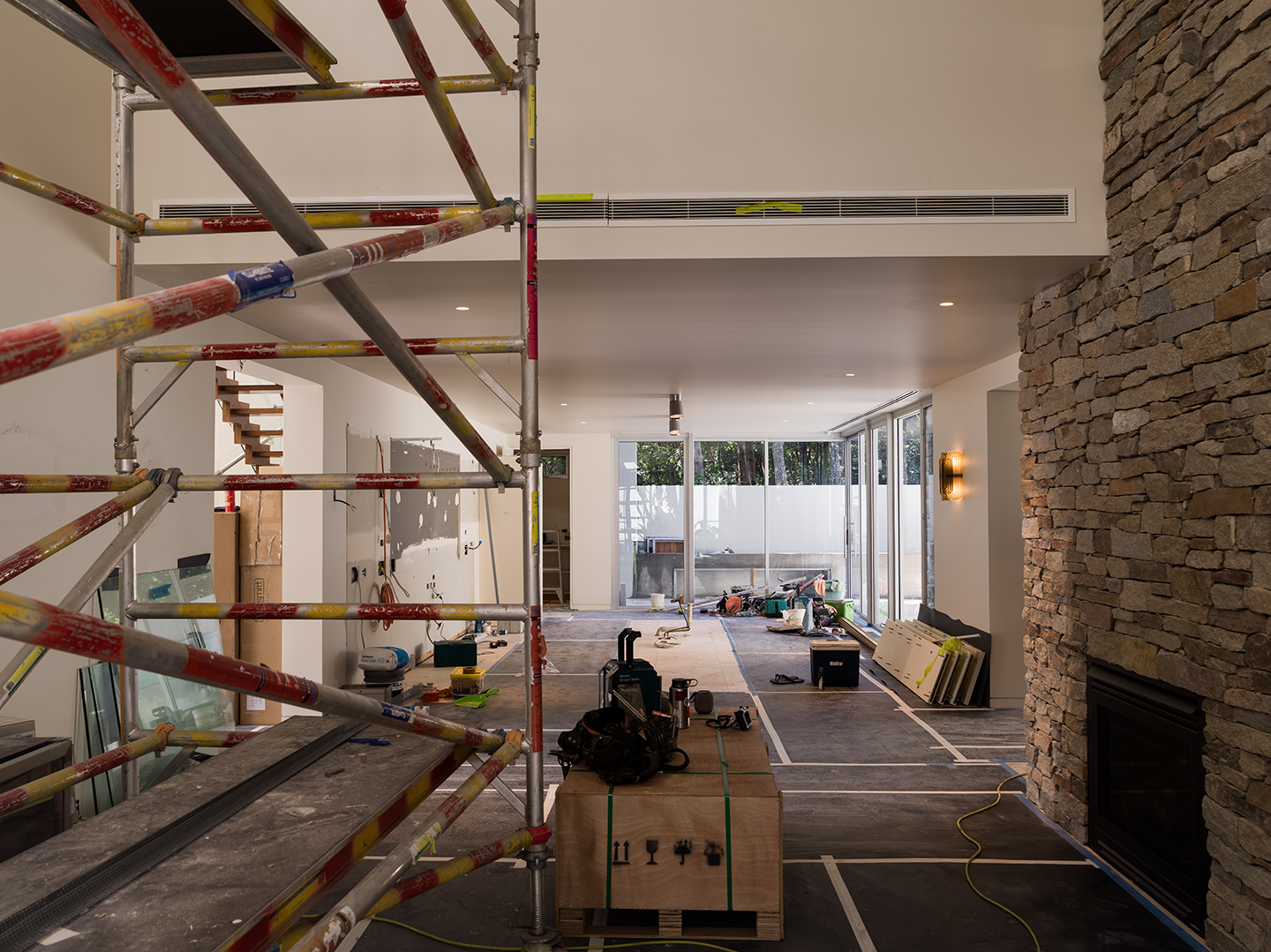 Interior of a house under renovation, with scaffolding, tools, and construction materials scattered around, large windows at the back, and a stone fireplace on the right.