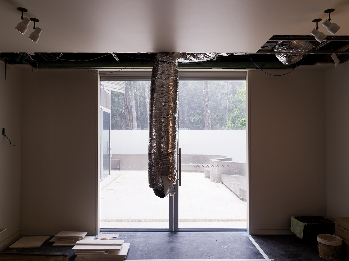 Interior of a room under renovation with an unfinished ceiling, exposed ductwork, and a large sliding glass door leading to an outdoor patio with trees.