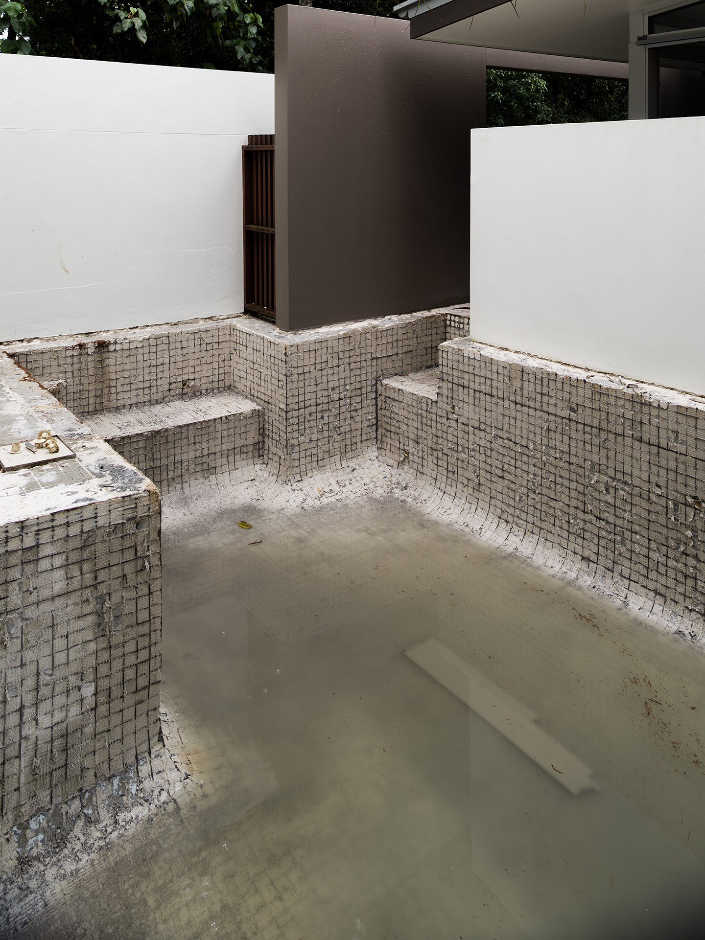 Empty swimming pool with dirty water, surrounded by white and gray walls, during construction or renovation.