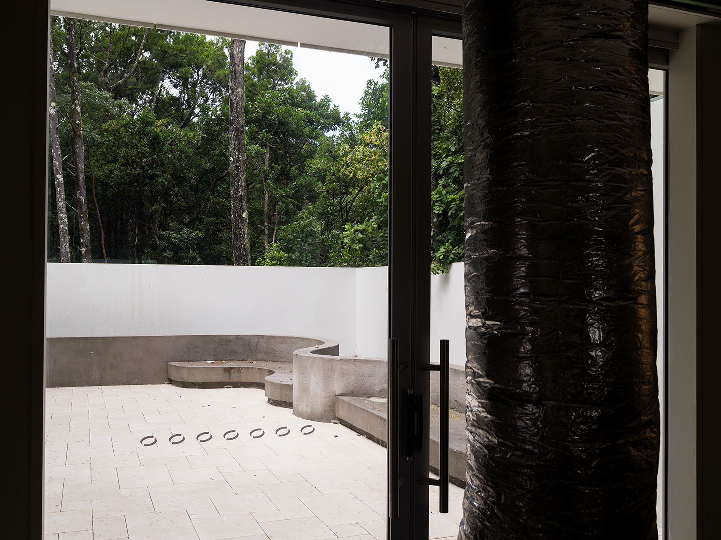 View from inside a modern building looking out onto a patio with concrete seating, enclosed by a white curved wall, surrounded by trees and greenery.