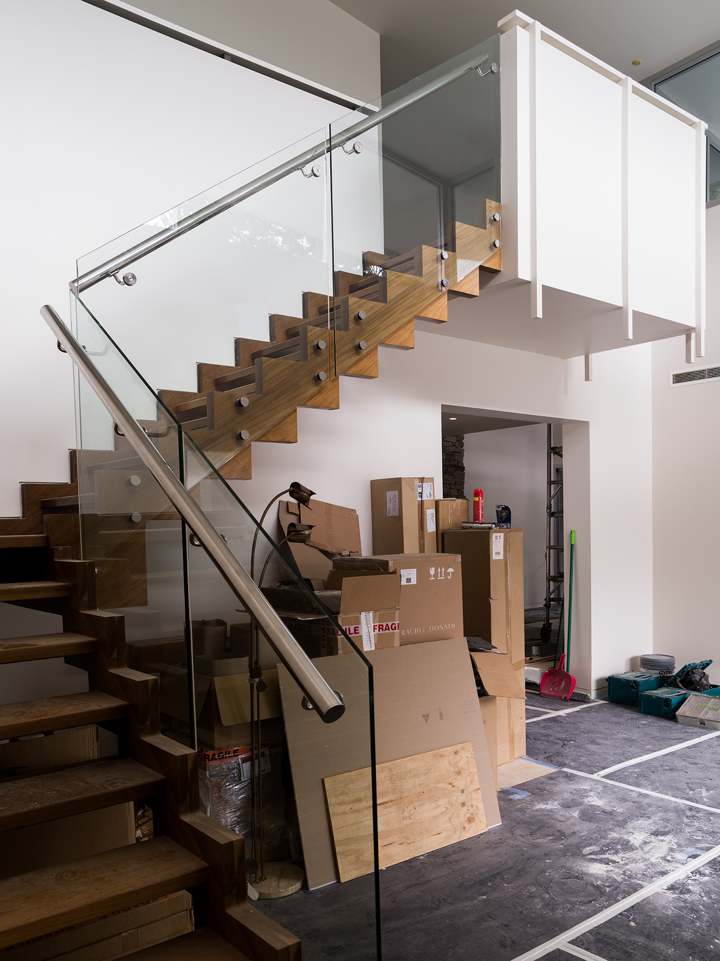 Interior view of a modern house under construction with a wooden staircase, glass railing, and boxes of supplies.