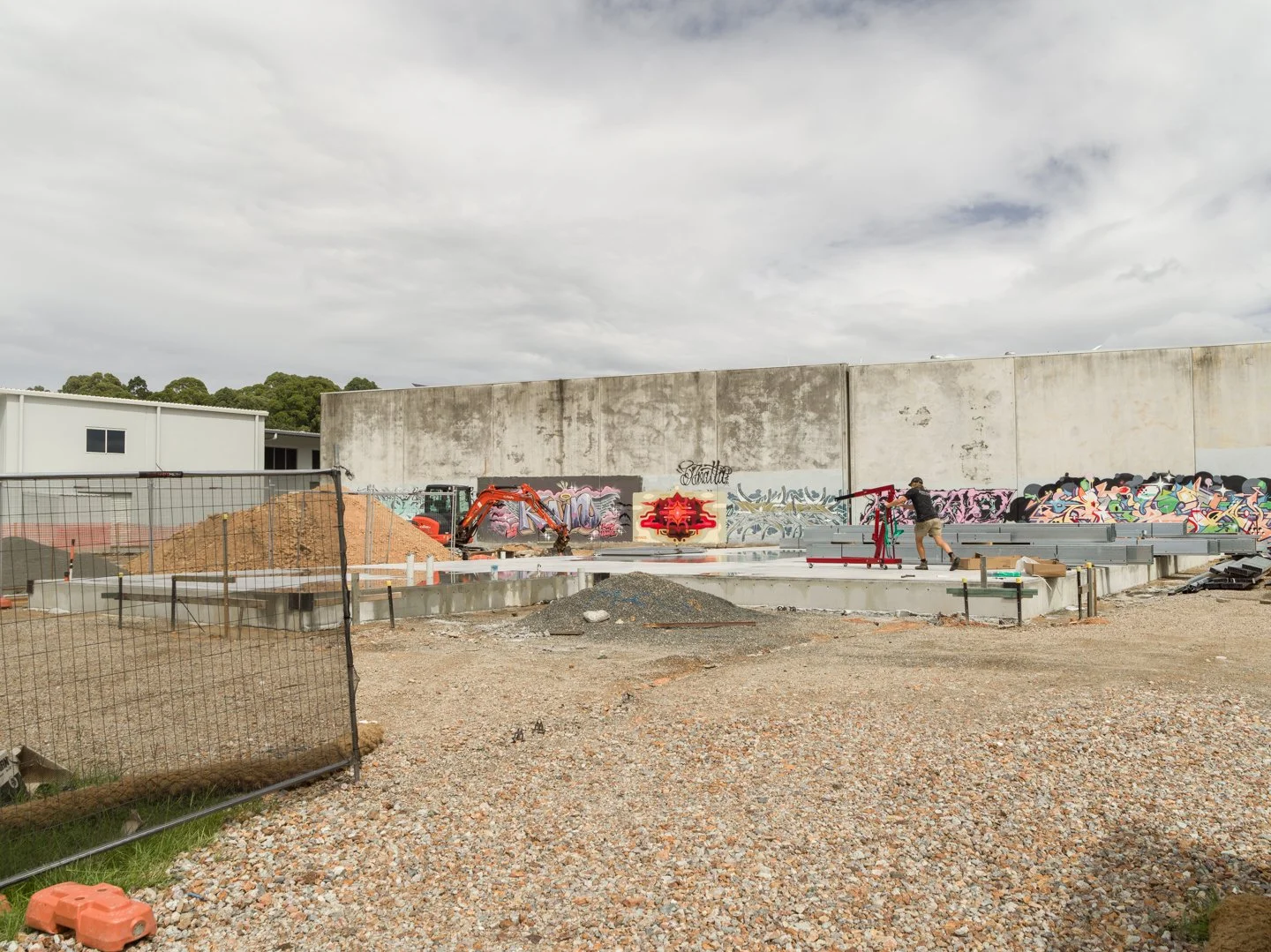 Construction site with a worker and graffiti-covered concrete wall in the background, gravel ground, construction equipment, and a small excavator.