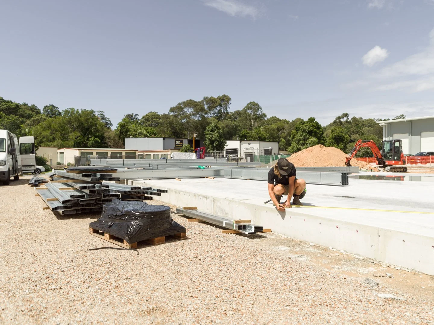 A construction worker crouched on the ground measuring a concrete slab with a tape measure at an outdoor construction site. Piles of metal beams, black plastic-wrapped items, a mini excavator, and trucks are visible in the background, with trees and 