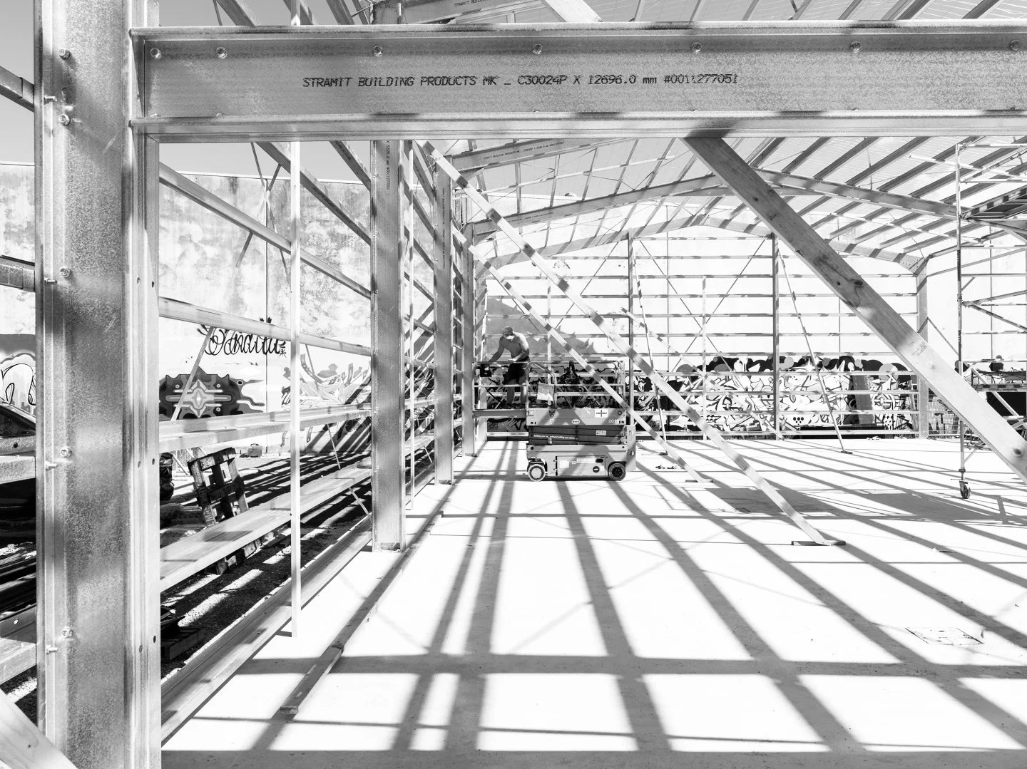 Construction site with metal framework and scaffolding, with a worker on a lift working on the structure. Graffiti is visible on the wall in the background, and sunlight creates shadows on the ground.