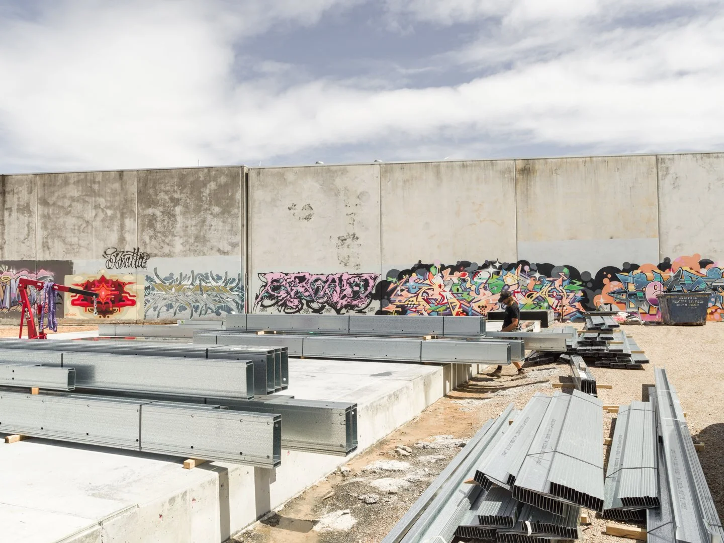 Construction site with a concrete wall covered in colorful graffiti art, metal beams, and construction materials, with a worker walking among the materials under a partly cloudy sky.