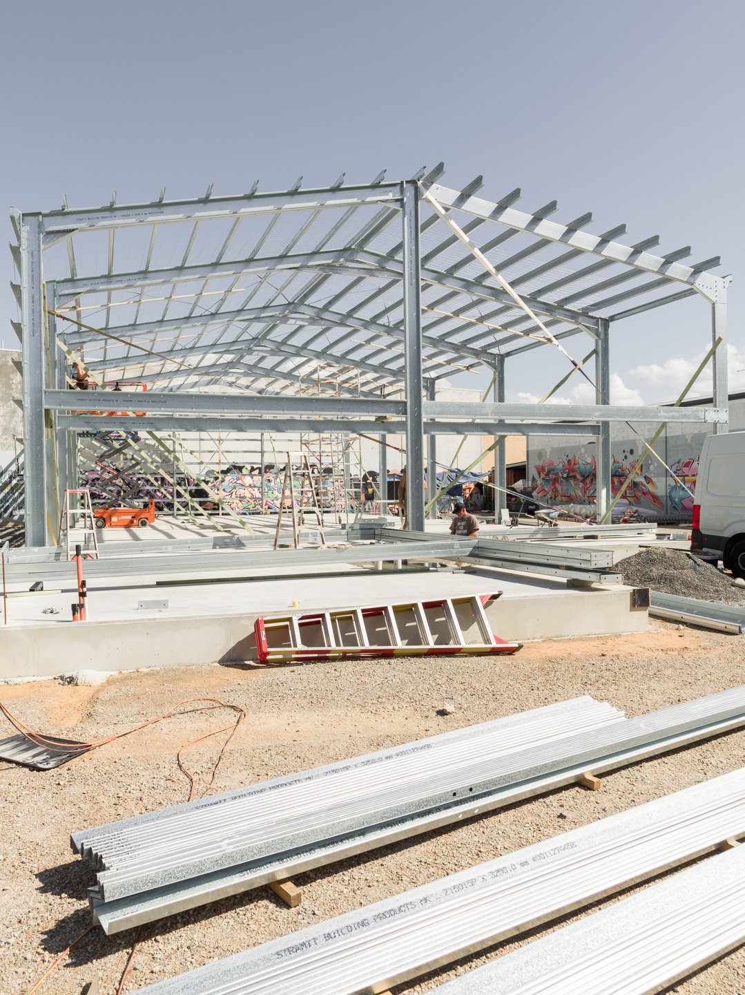 Steel frame building under construction with workers, ladders, and construction materials on the ground, with graffiti art on the background wall.