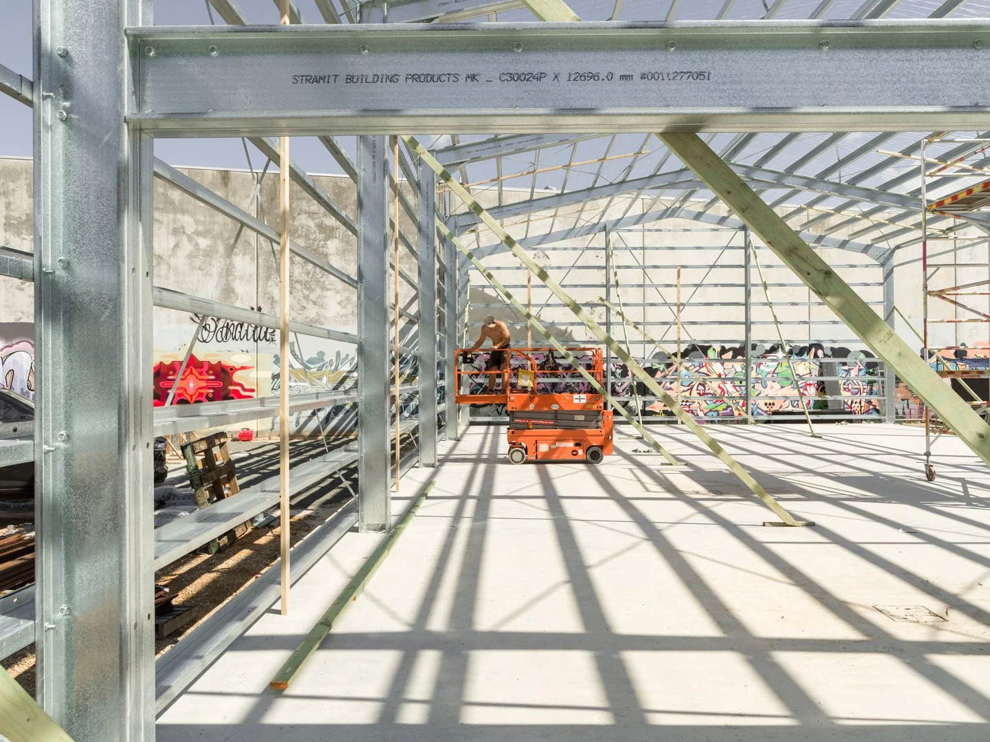 Construction worker on an orange lift working on a metal building frame with graffiti mural in the background.