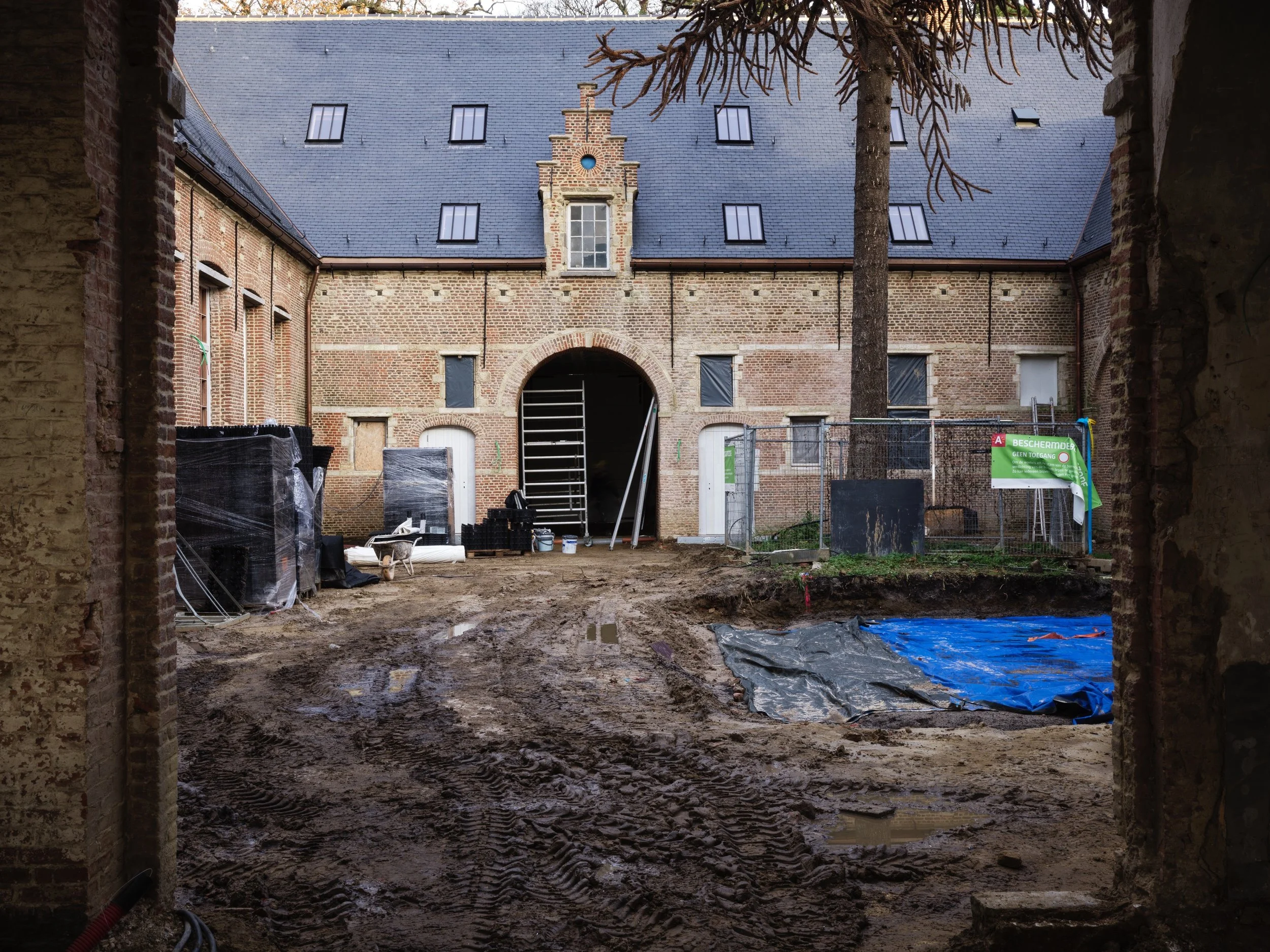 View of an old brick building under renovation seen from inside a construction site with muddy ground, construction materials, and fenced-off area.