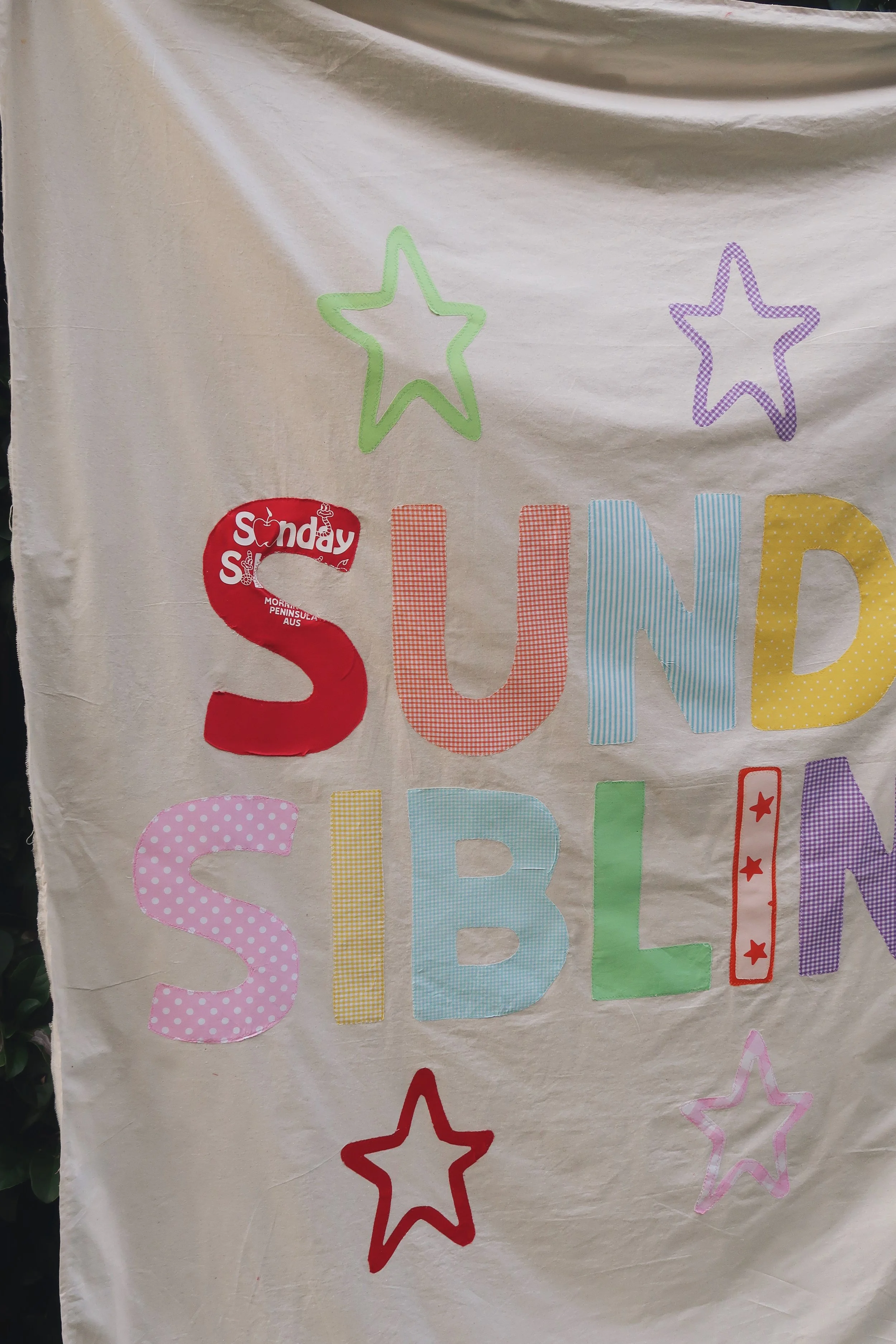 A colorful, decorative fabric with the word 'SUNDAY' in large, multicolored letters, surrounded by outlined star shapes in green, purple, red, pink, blue, and yellow, and small red stars inside a rectangle.