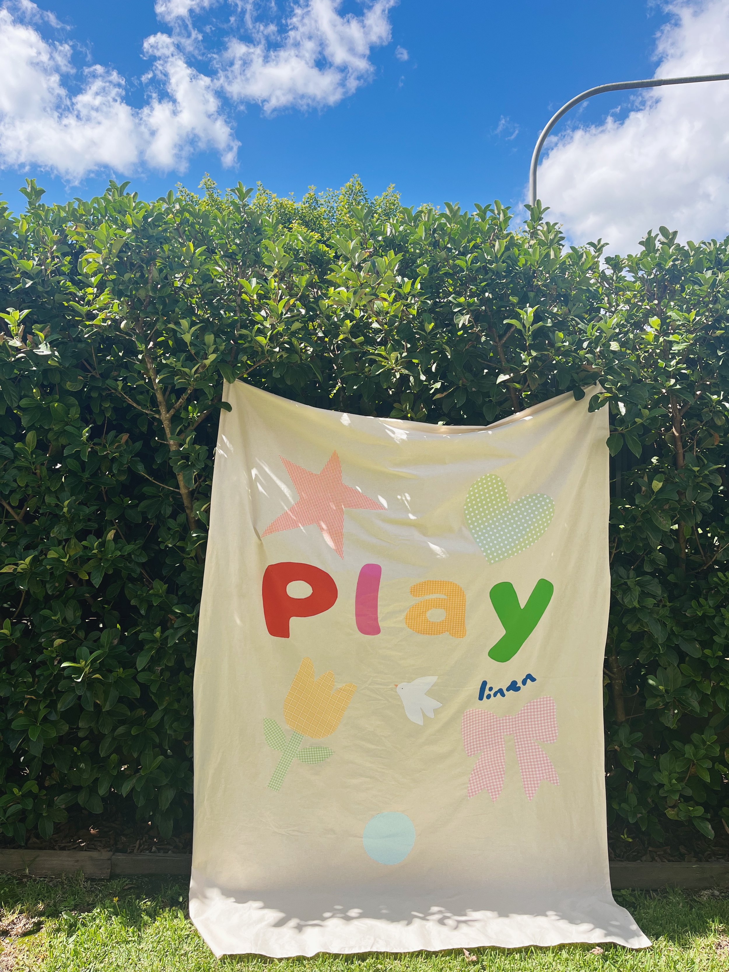 Colorful banner with the word 'play' in multicolored letters, decorated with stars, hearts, and other shapes, hanging outdoors in front of green bushes and a bright blue sky with white clouds.