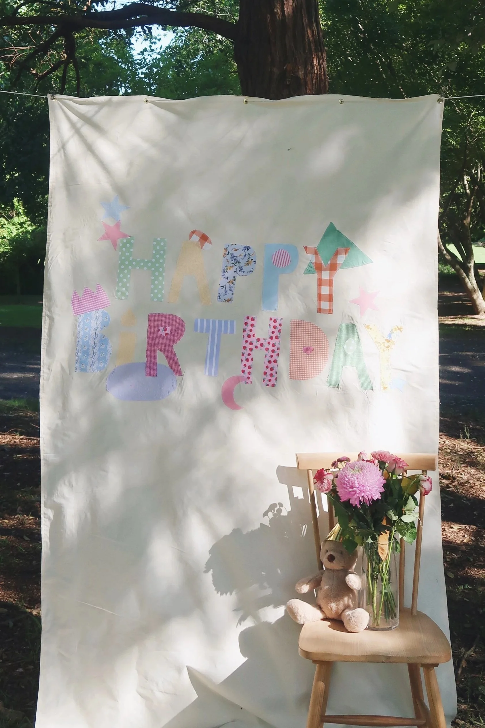 A white fabric backdrop with multicolored cut-out letters spelling 'HAPPY BIRTHDAY' hangs in an outdoor setting among trees. In front of the backdrop, a small wooden chair holds a glass vase with pink flowers and a plush teddy bear against a backdrop of green foliage and sunlight filtering through the trees.