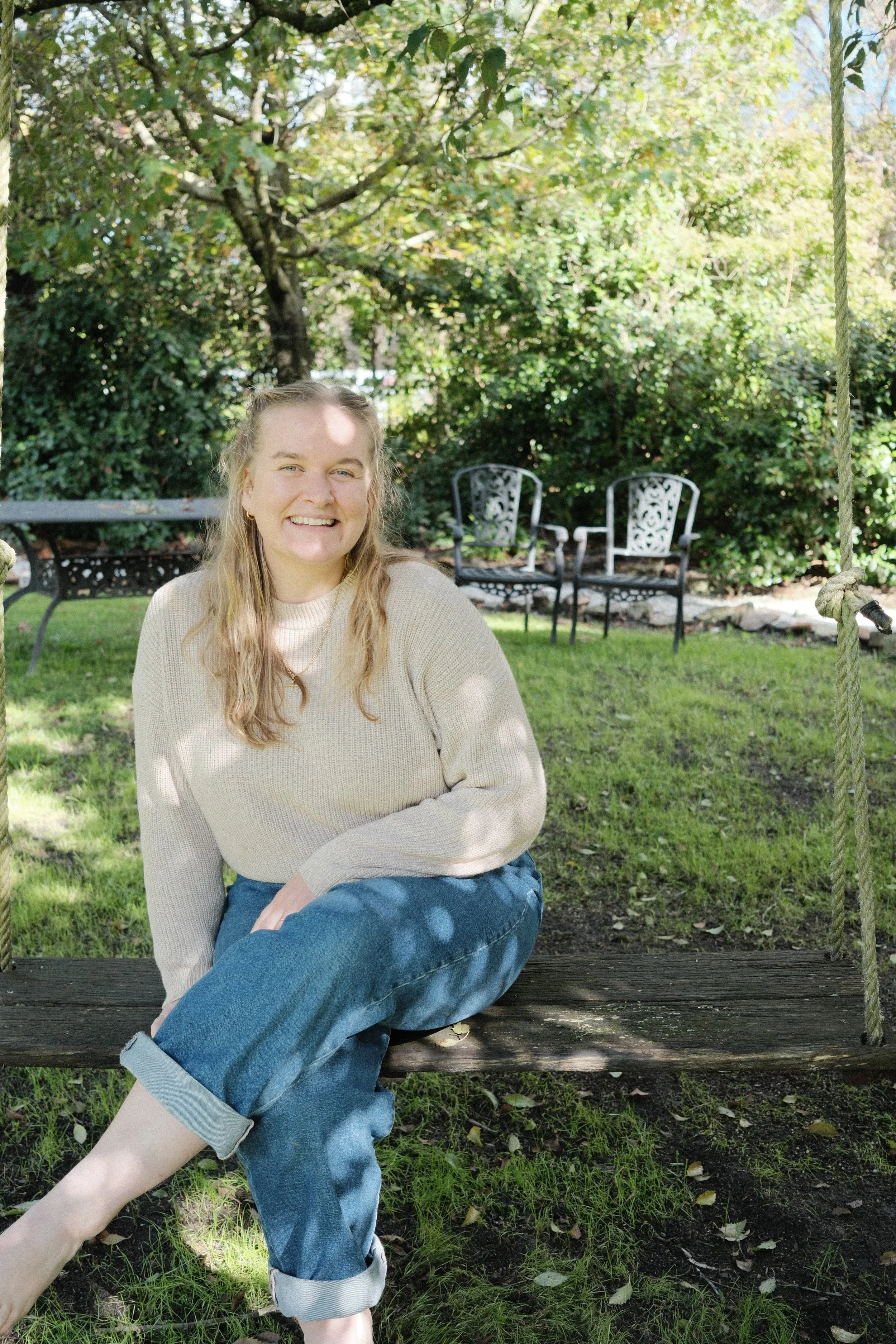 A young woman sitting on a wooden bench with crossed legs, smiling, in a backyard with green grass, bushes, and trees, and two metal chairs in the background.