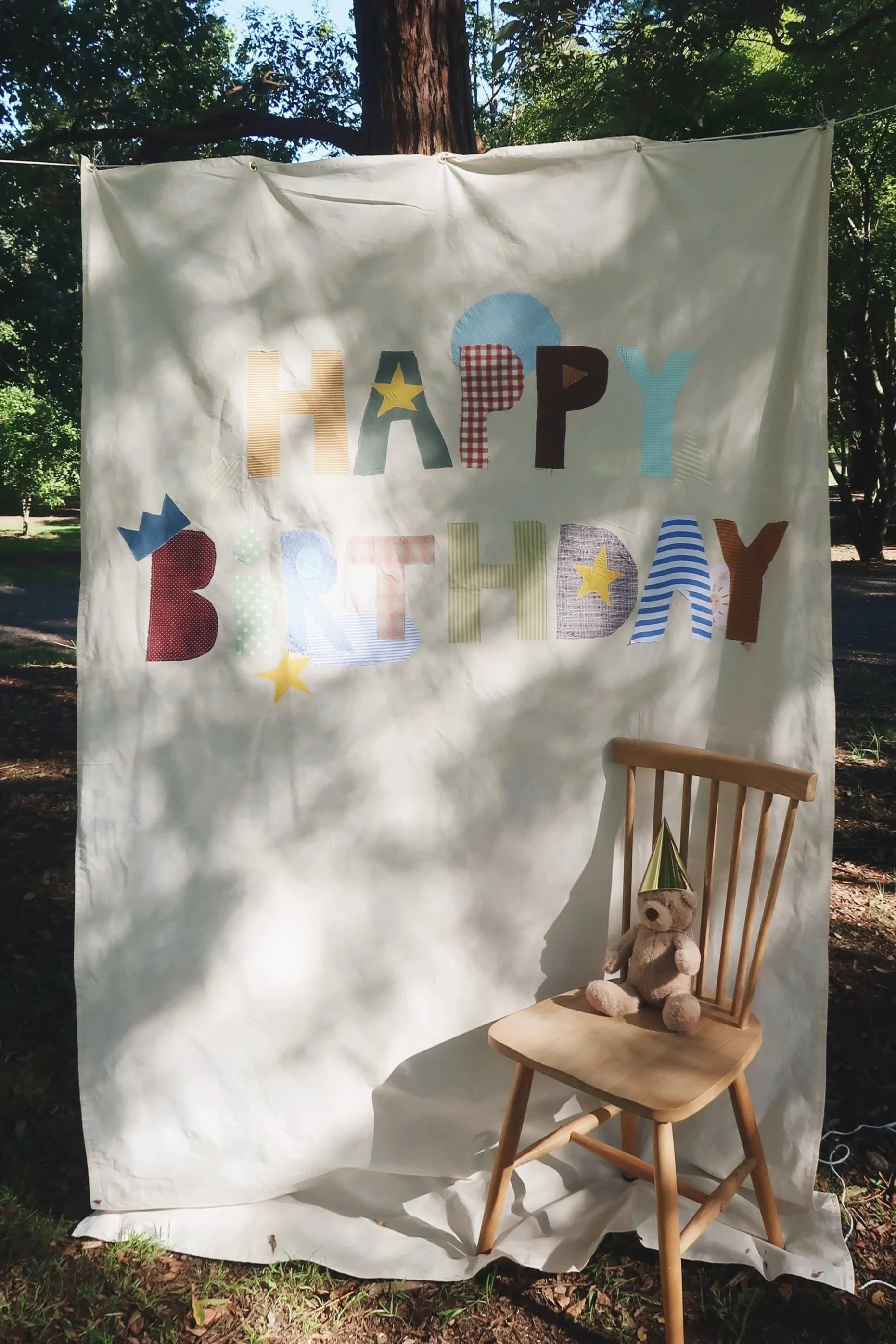 Decorative banner with the words 'HAPPY BIRTHDAY' in colorful letters, hanging outdoors with trees in the background, and a wooden chair with a teddy bear wearing a party hat sitting on it.