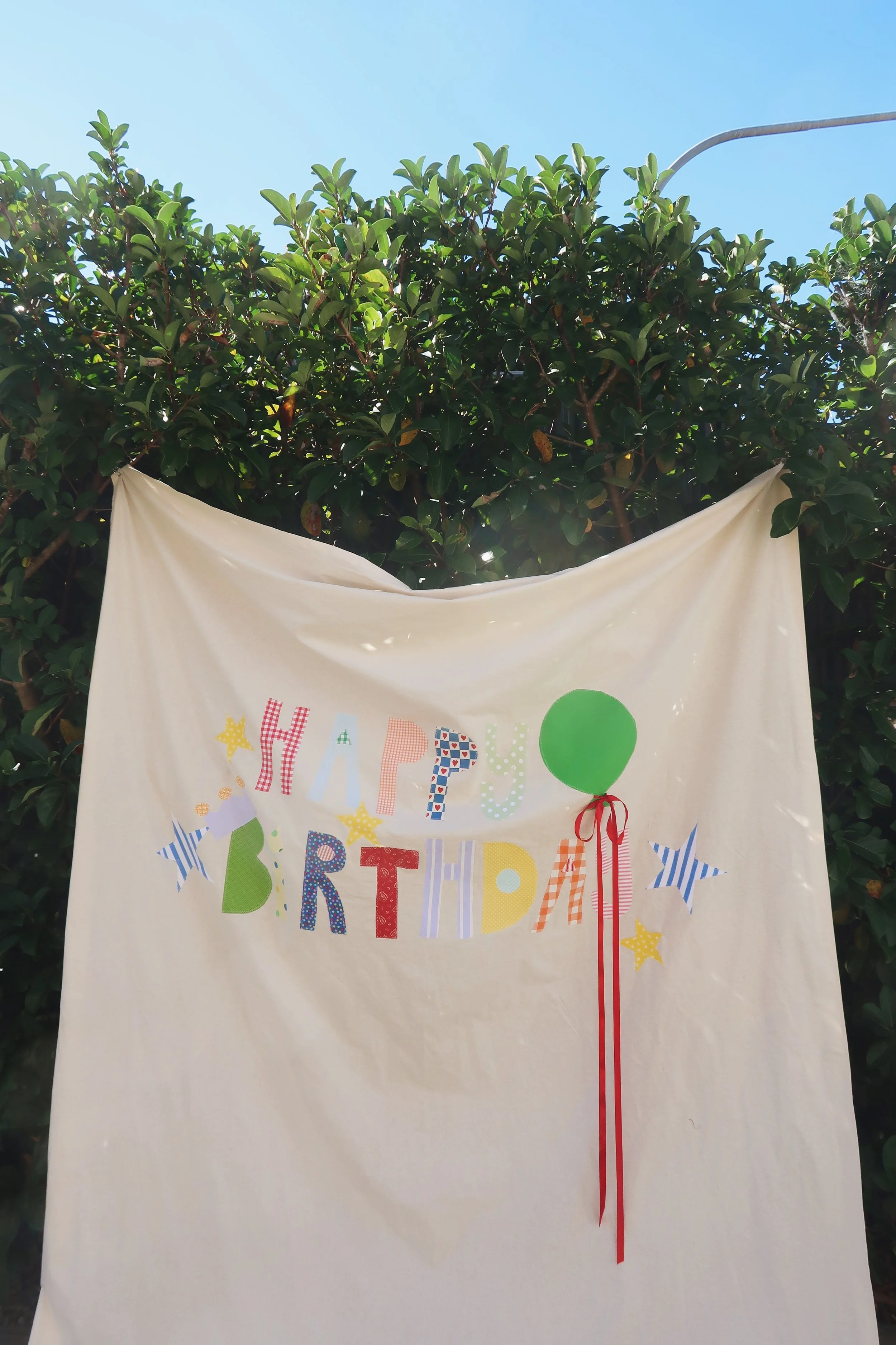 A colorful birthday banner with the words "Happy Birthday" and decorative stars, hearts, and a balloon, hanging in front of a green hedge under a clear blue sky.