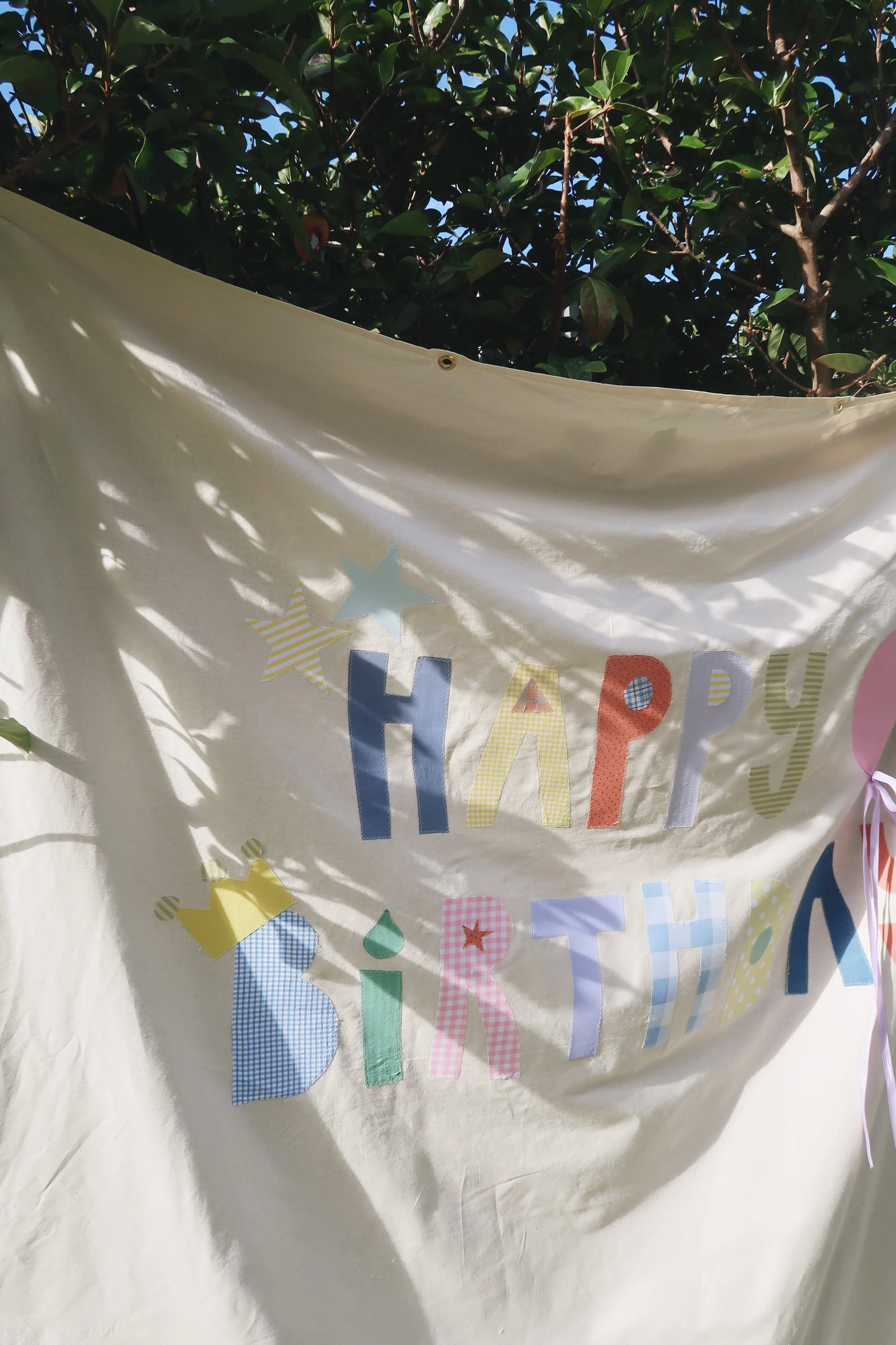Part of a birthday banner with colorful alphabet letters and star cutouts hanging on a white fabric, with tree branches and leaves in the background.