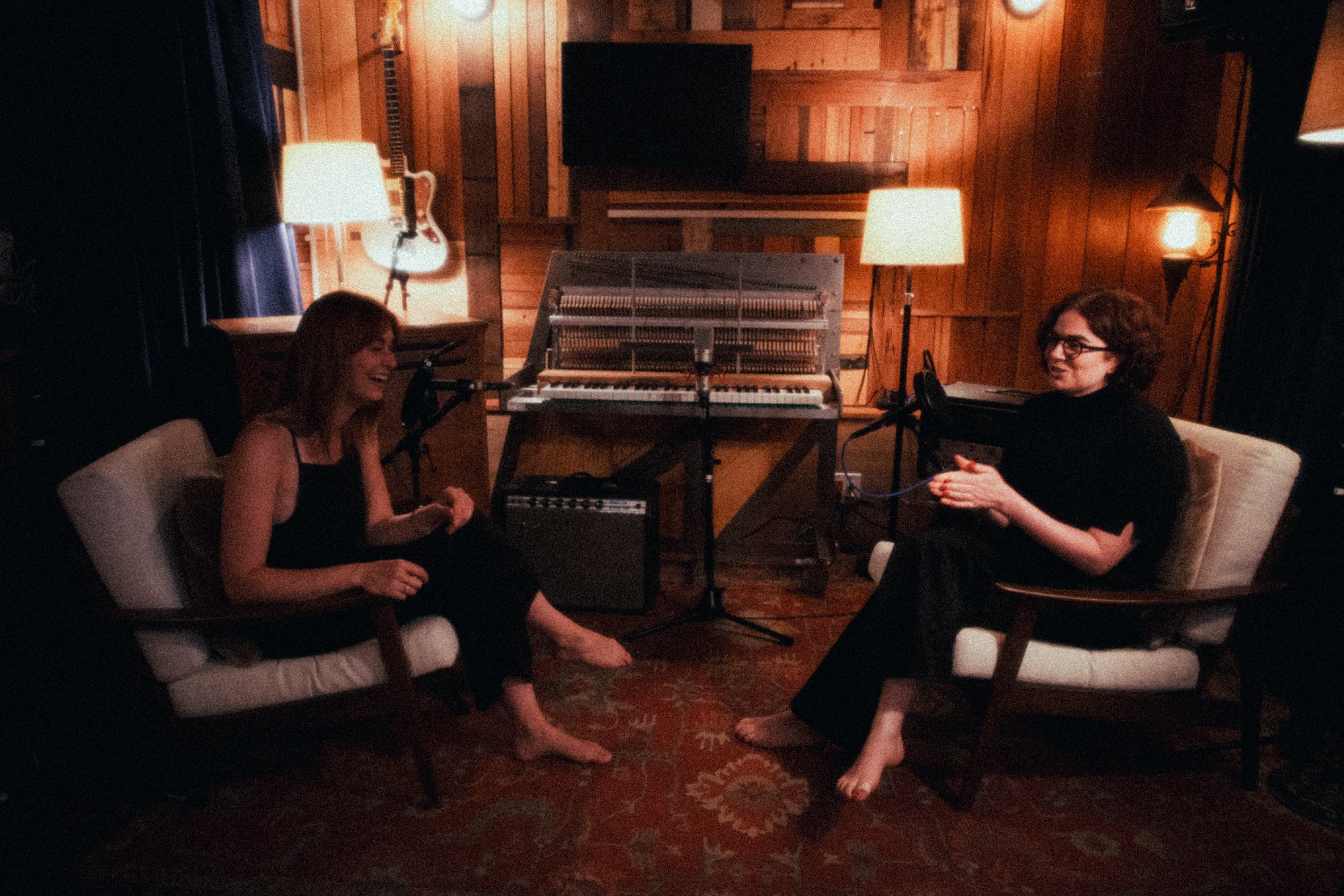 Two women are seated in a warmly lit recording studio with wooden walls and flooring. The woman on the left is laughing, has red hair, and wears a black sleeveless top. The woman on the right has short, curly brown hair, wears glasses, and is looking towards the camera. Both are barefoot and sitting on upholstered chairs. In between them are microphones, and behind them a vintage upright piano with an open top exposing the strings and hammers. The studio has various musical instruments and equipment, including a guitar hanging on the wall and several lamps providing soft lighting.