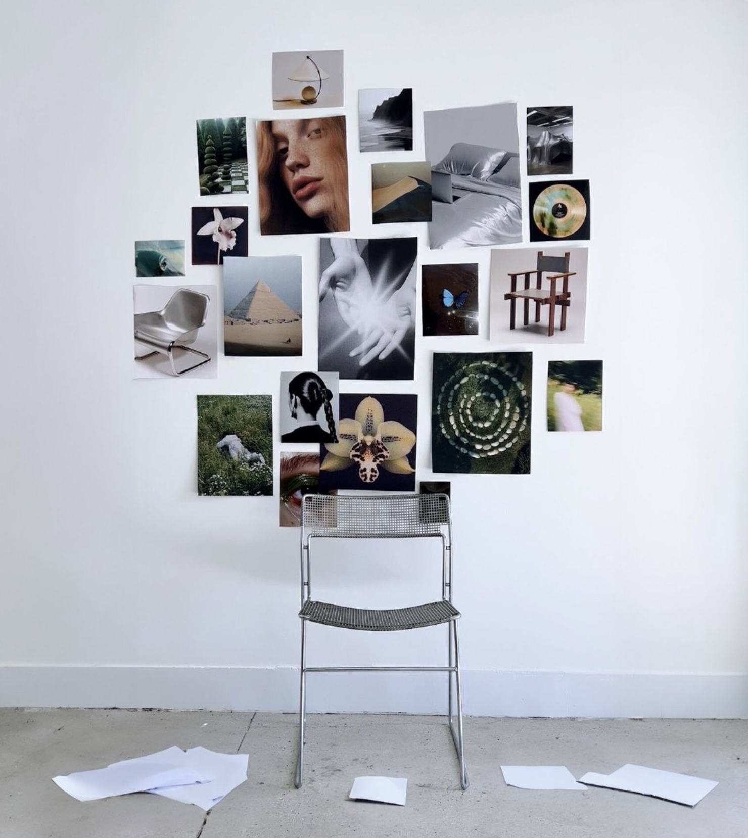 A wall with various photographs arranged in a collage, including portraits, objects, landscapes, and abstract images. In front of the wall is a metal chair, and on the floor are scattered sheets of paper.