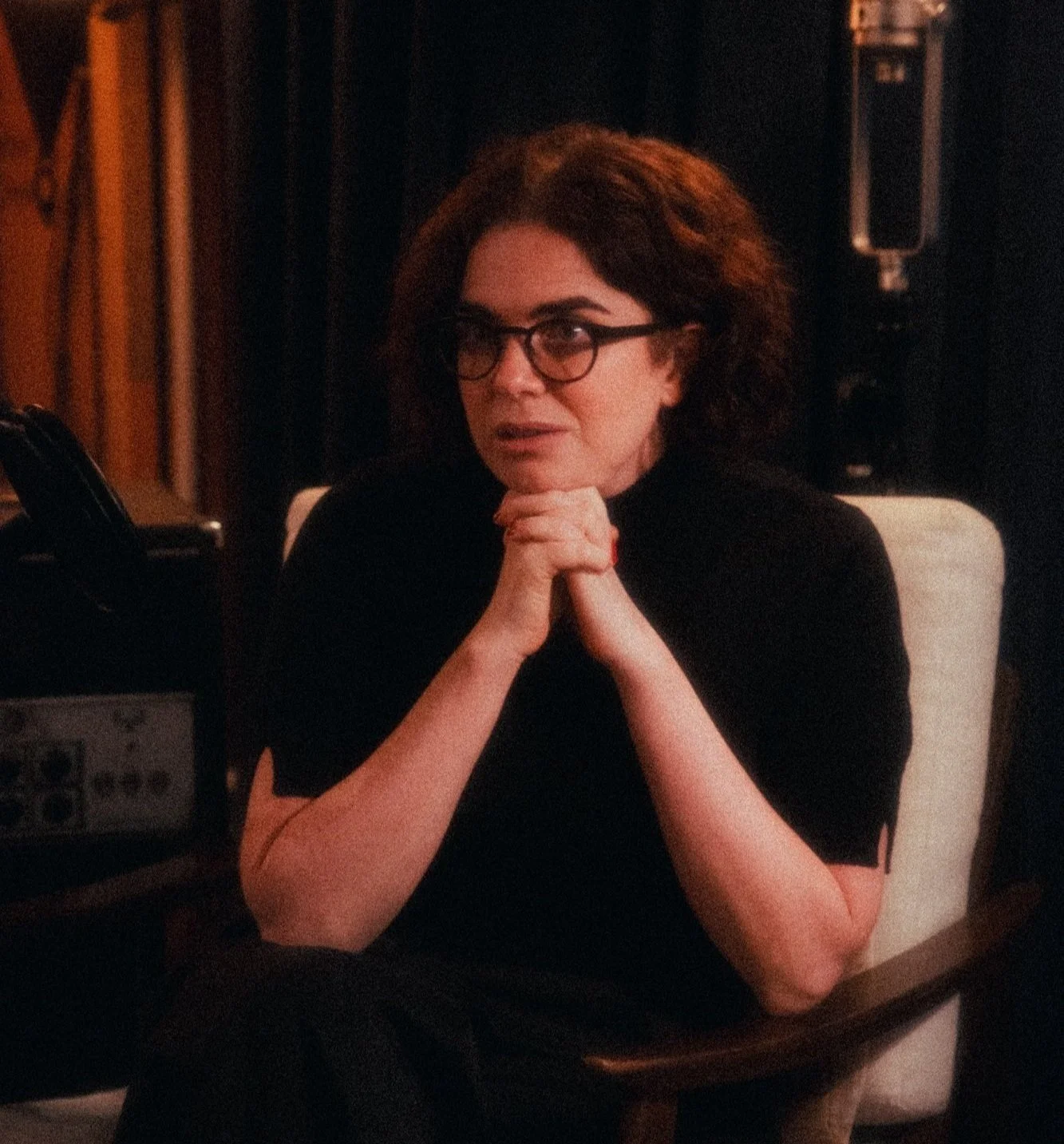 A woman with black hair and glasses, wearing a black top, sitting on a white chair with her hands clasped under her chin.