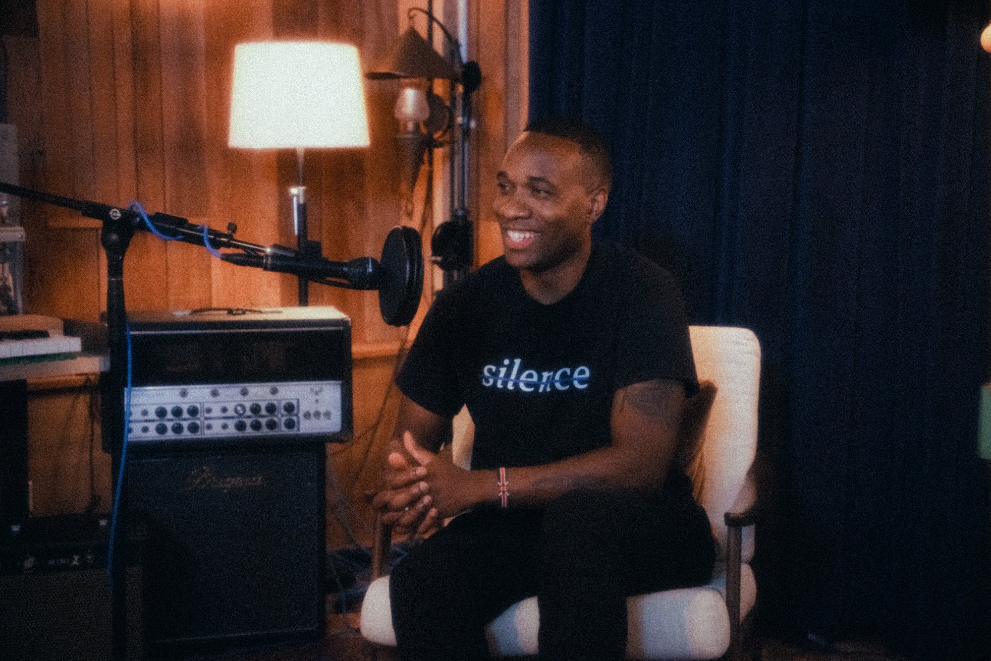 A smiling person sitting in a white chair in a recording studio, wearing a black T-shirt with the word 'silence' printed on it. There is a microphone and audio equipment on a stand to the left, and a lamp providing warm lighting in the background.