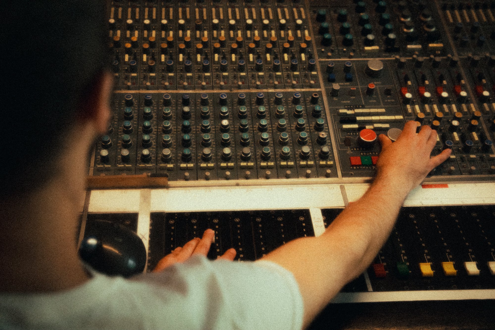Person operating a large audio mixing console with numerous knobs, sliders, and buttons in a recording studio.