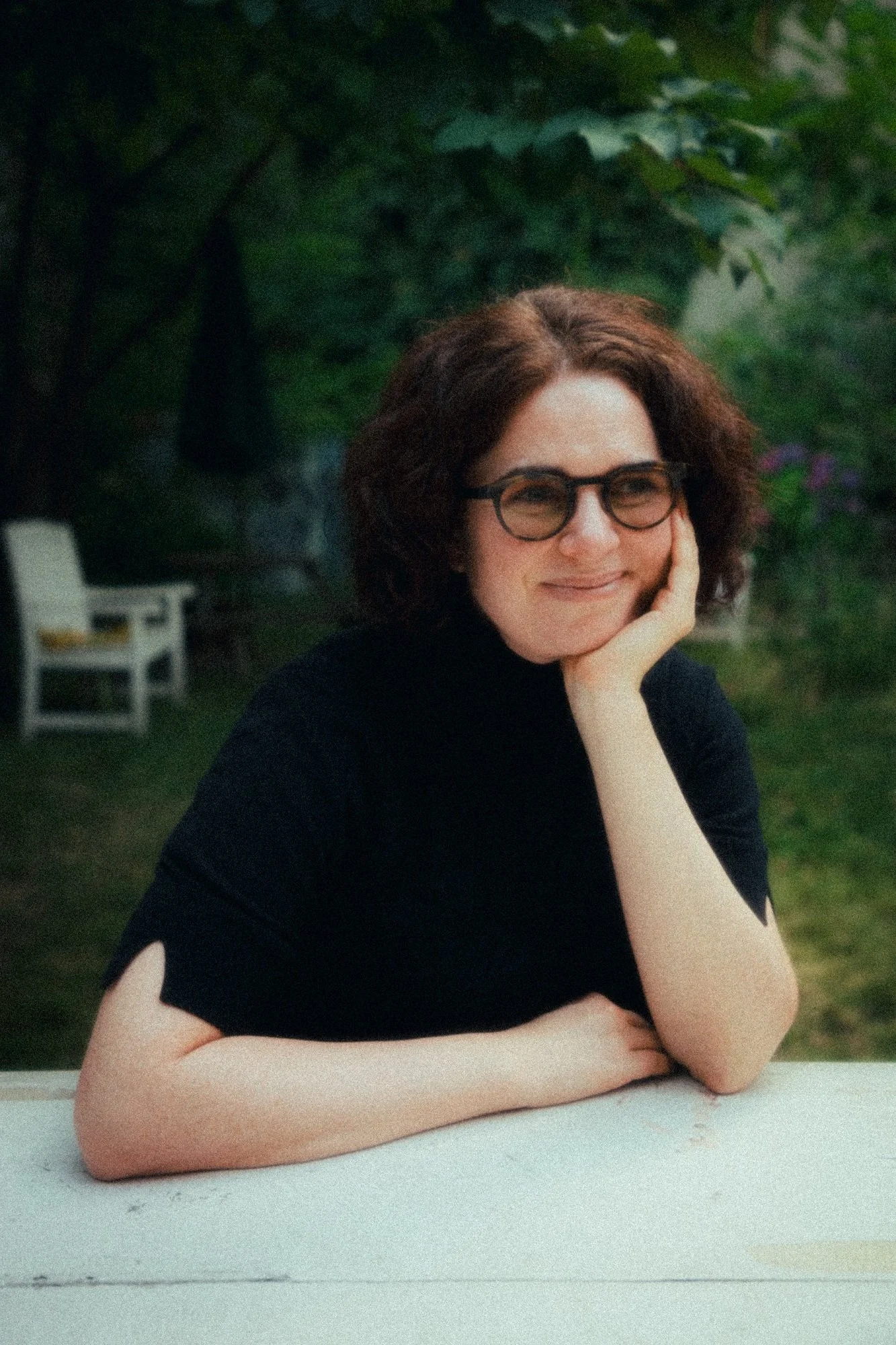 A smiling woman with short curly brown hair and glasses, sitting outdoors at a table with her chin resting on her hand, in a garden with greenery and a white bench in the background.