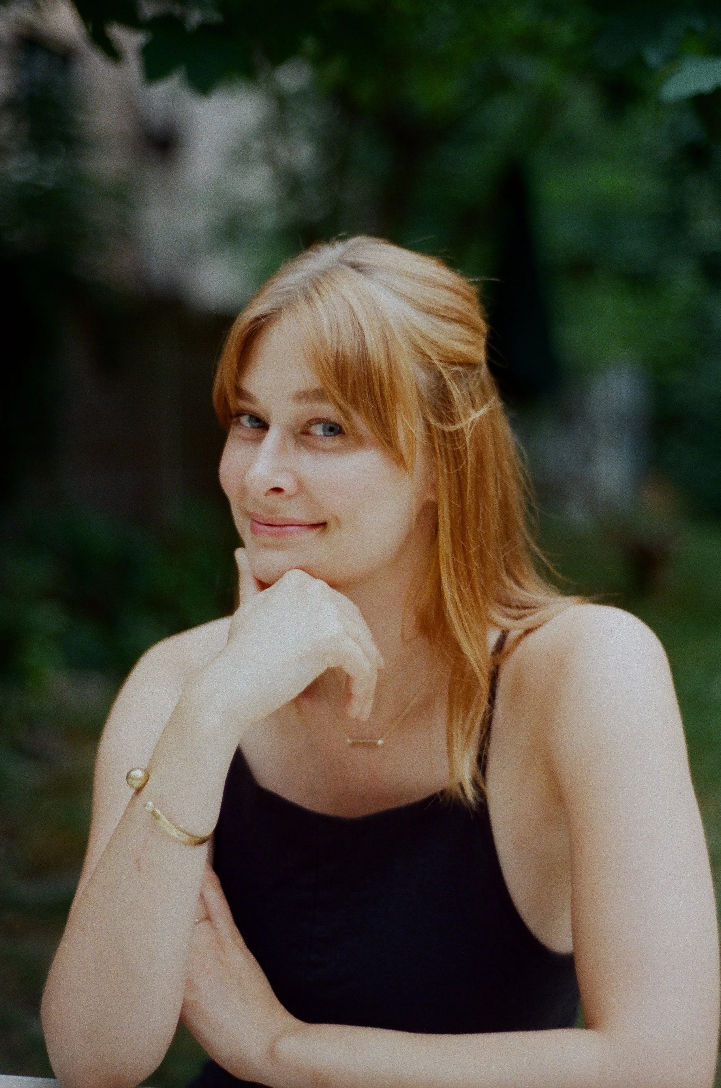 A young woman with red hair and blue eyes smiling with her hand resting on her chin, outdoors with greenery in the background.