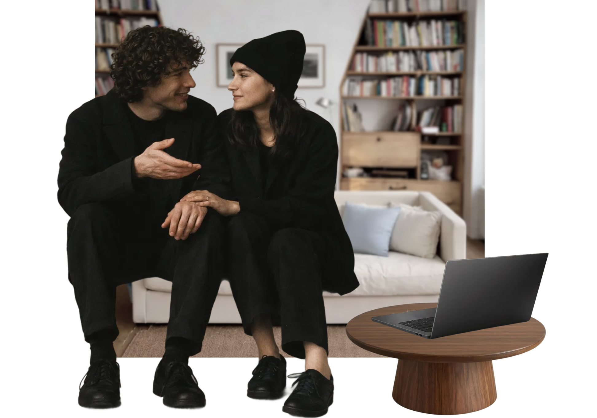 Two people sitting on a white sofa, engaged in conversation, with a laptop on a round wooden table in front of them. The background features bookshelves filled with books.