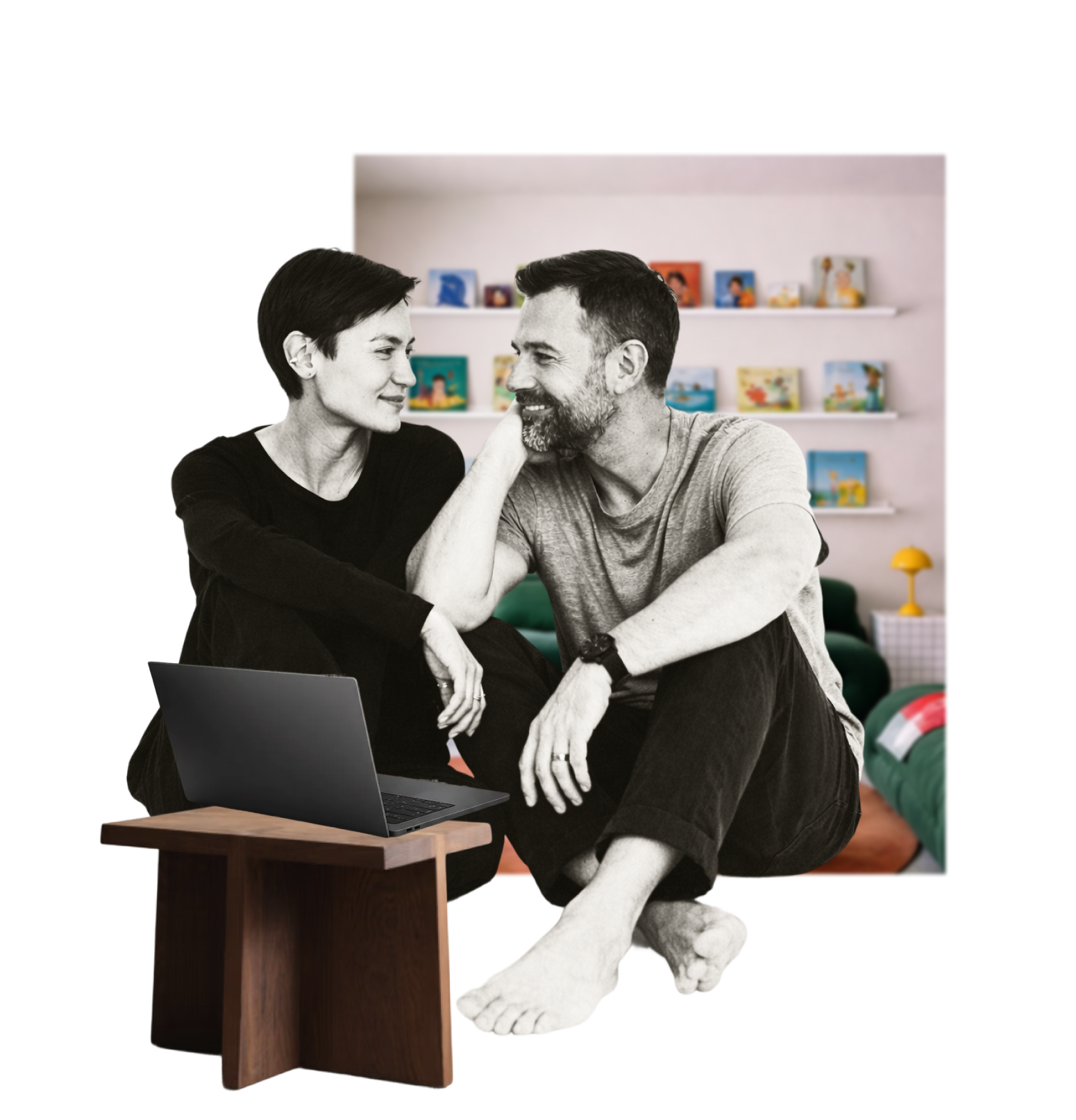 A black-and-white photo of a woman and man sitting close together in a colorful room, with the woman smiling and looking at the man. A laptop is on a small wooden table in front of them, and the background features shelves with children's books and toys.
