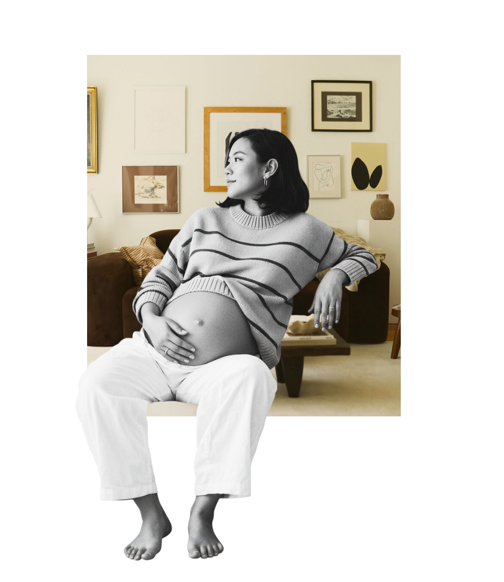 A pregnant woman sitting in a living room, cradling her belly, with framed artwork on the wall behind her.