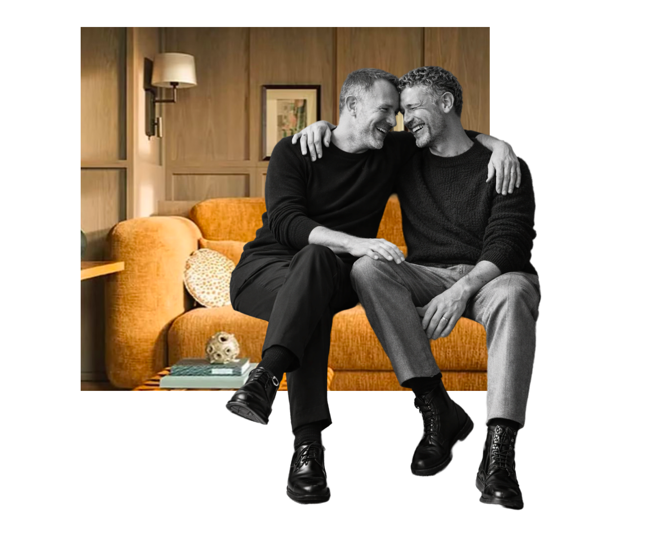 Two men embracing and smiling under warm lighting in a cozy, wood-paneled room with framed pictures on the wall. They are being interviewed about their love journey together.