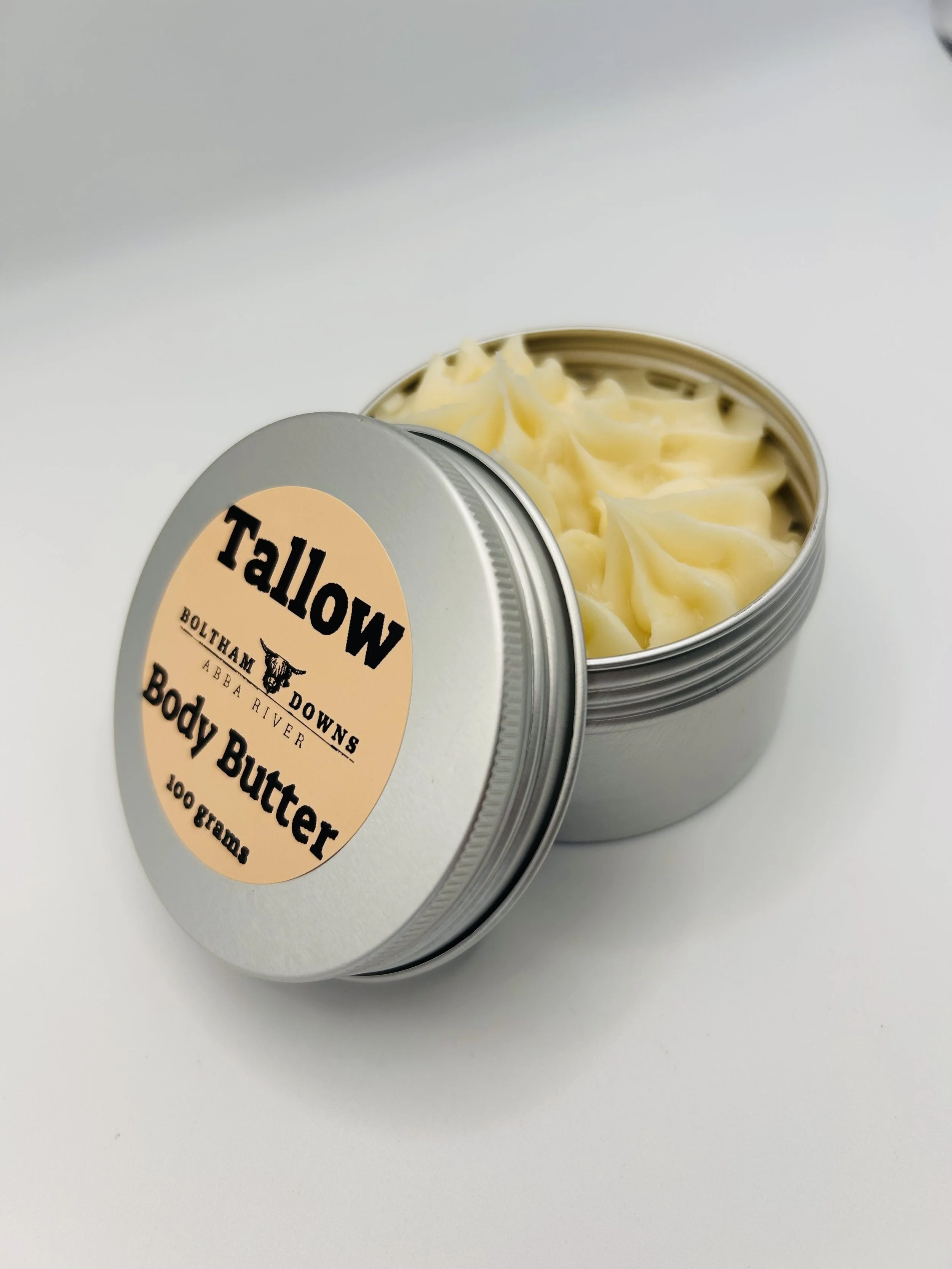 Open tin of Highland Park body butter with its lid tilted, revealing the creamy yellow substance inside on a white background.