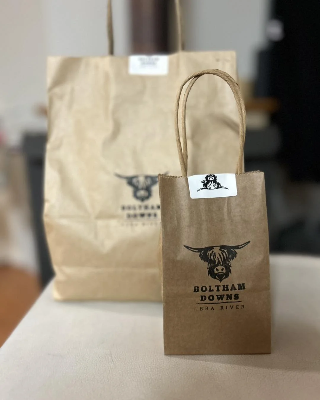 Two Baltham Downs BBQ River paper bags with a highland cow logo, one small and one large, placed on a table.