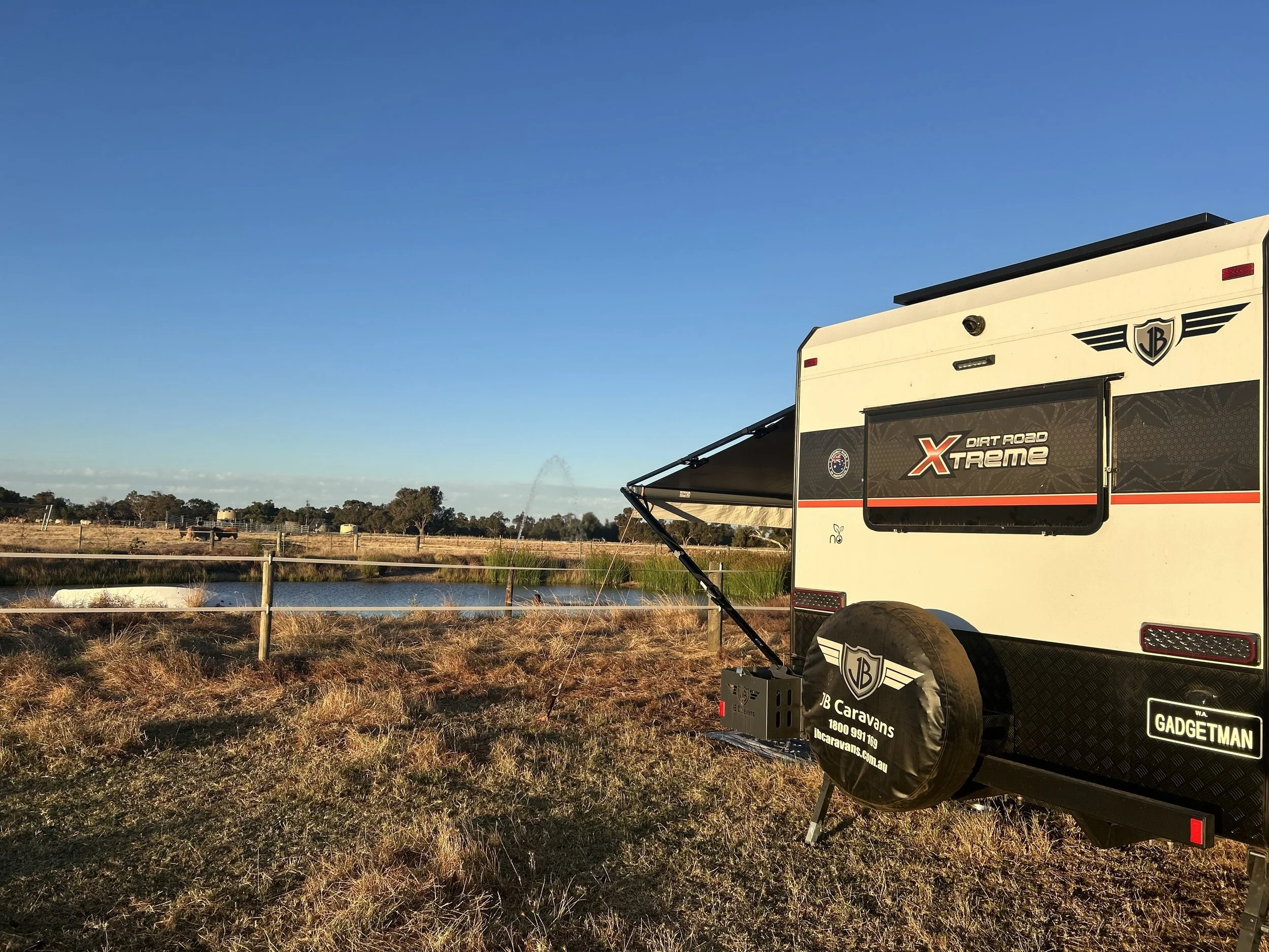Part of a travel trailer with a logo reading 'Gadgetman' and a window displaying 'X Dirt Road Xtreme'; the trailer is set against a natural landscape with grass, a small water body, and trees in the background. The sky above is clear with a few clouds.